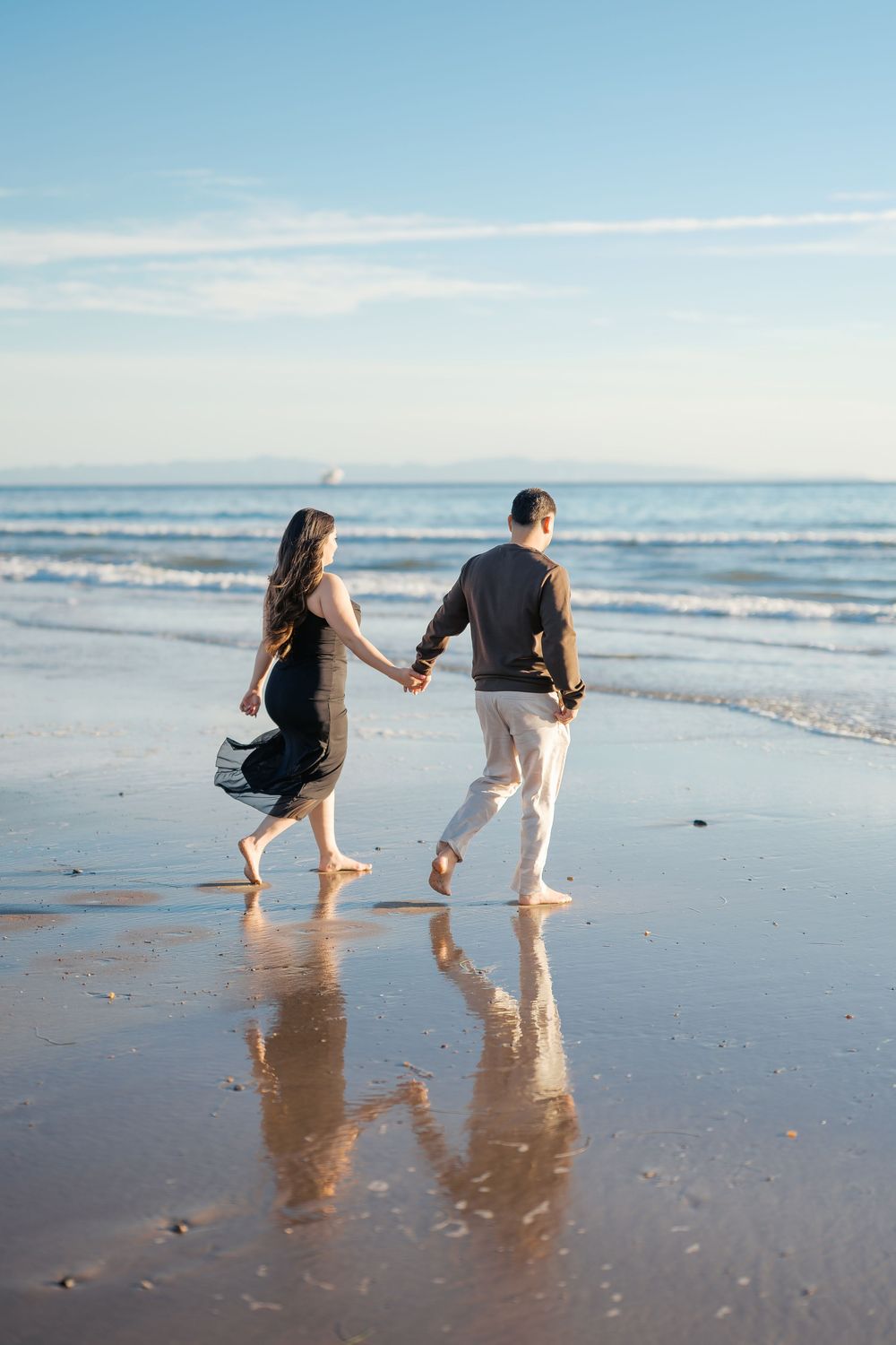 A couple walks hand in hand along a serene beach shoreline during golden hour, their reflections mirrored in the wet sand.