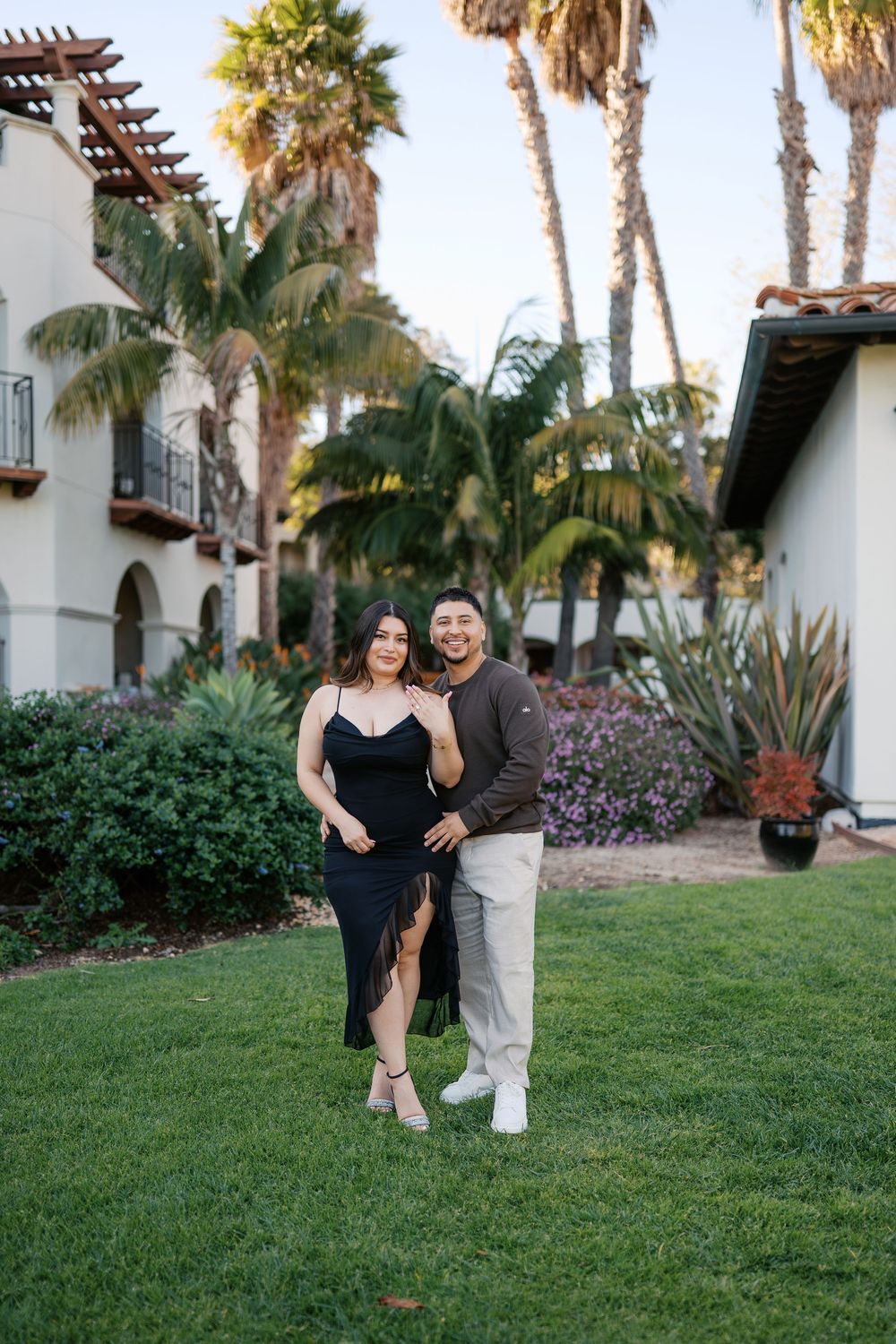 A couple poses playfully on a grassy lawn with palm trees and Spanish-style architecture in the background.