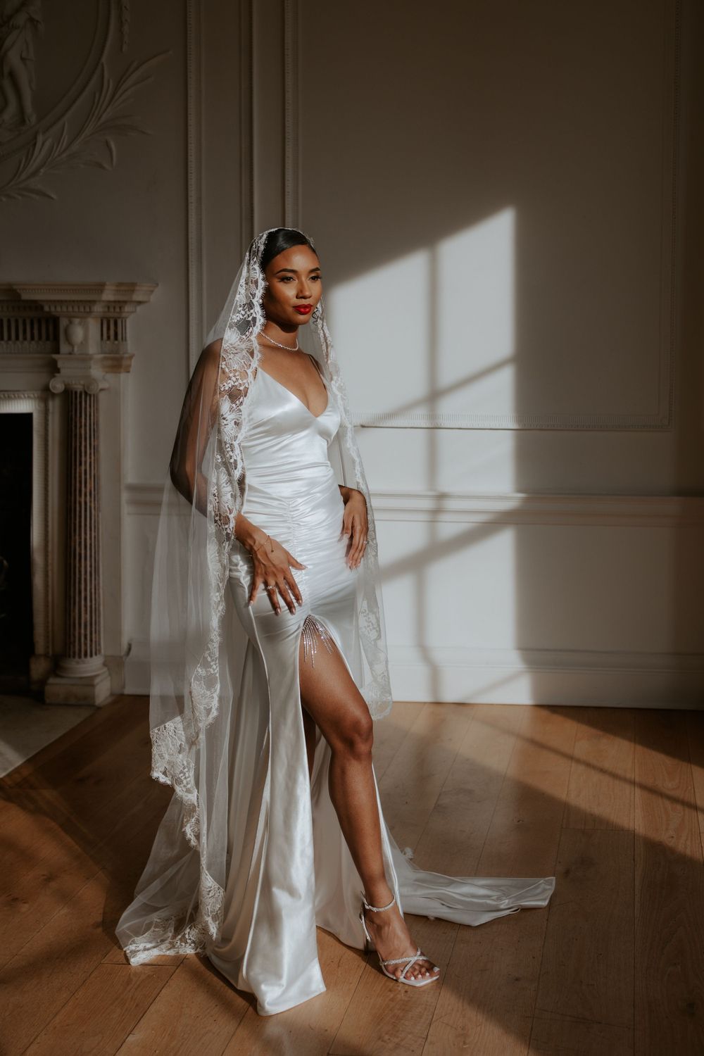 Elegant bride in white silk dress with lace veil poses in sunlit room with classic architectural details.