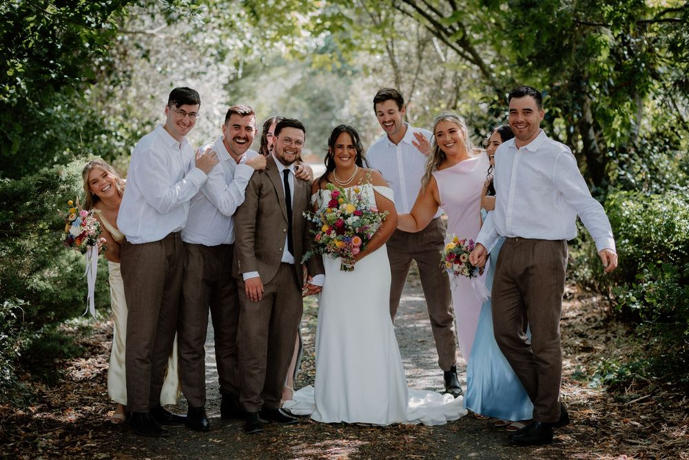 A wedding party in white and brown attire poses together on a tree-lined path during an outdoor ceremony.