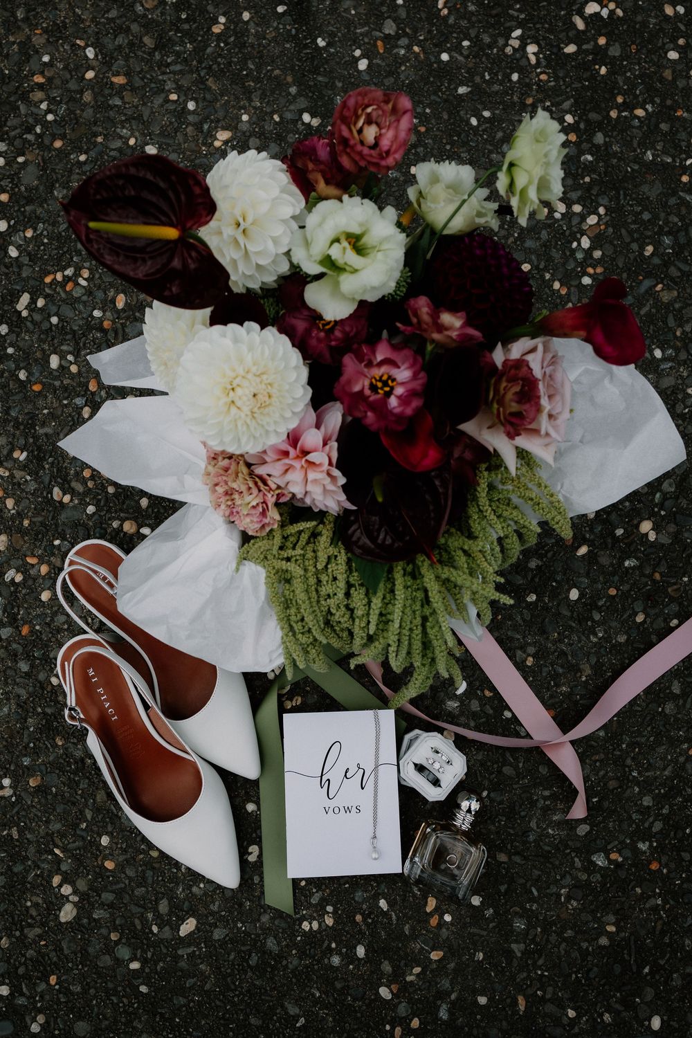 Wedding details flatlay with white shoes, burgundy flowers, and handwritten note card on dark textured surface.