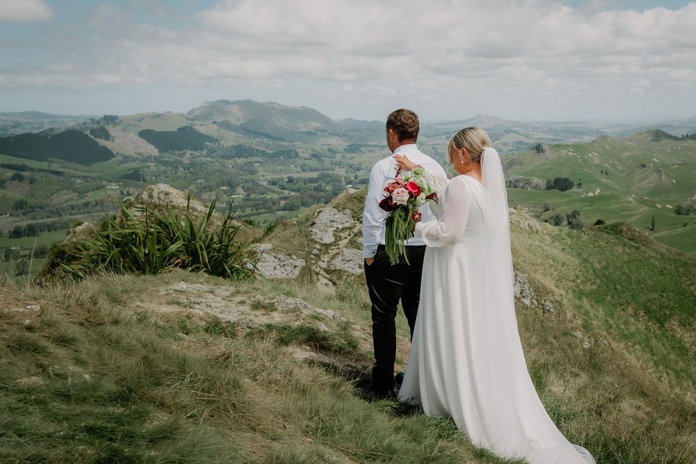 A bride and groom stand together on a hilltop overlooking rolling green mountains on their wedding day.