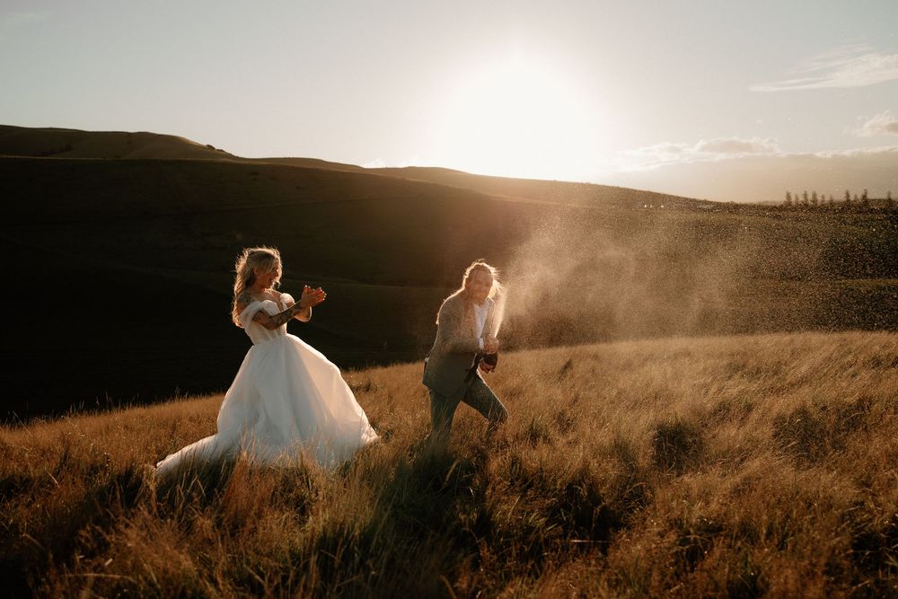 Couple in wedding attire walking through golden field at sunset with mountains in background.