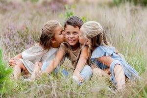 drie kinderen zittend op de grond. Tweeling zusjes geven hun broer tegelijk een kus op de wang.