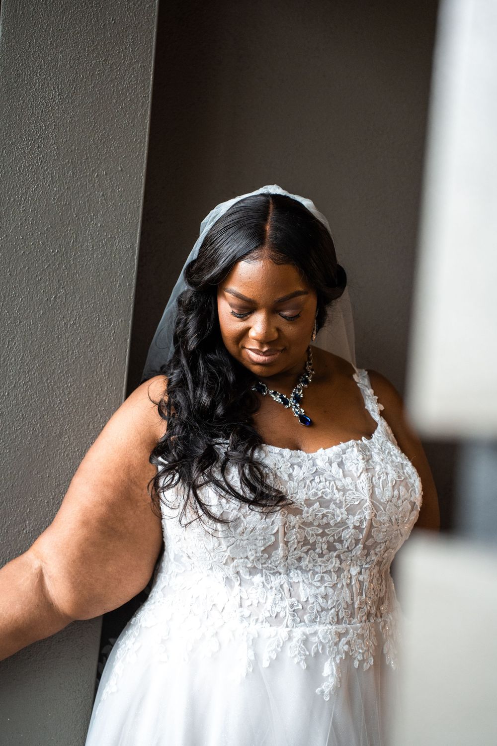A bride wearing a white lace wedding gown with long wavy hair and elegant necklace looks down with a gentle smile at Carollwood Country Club