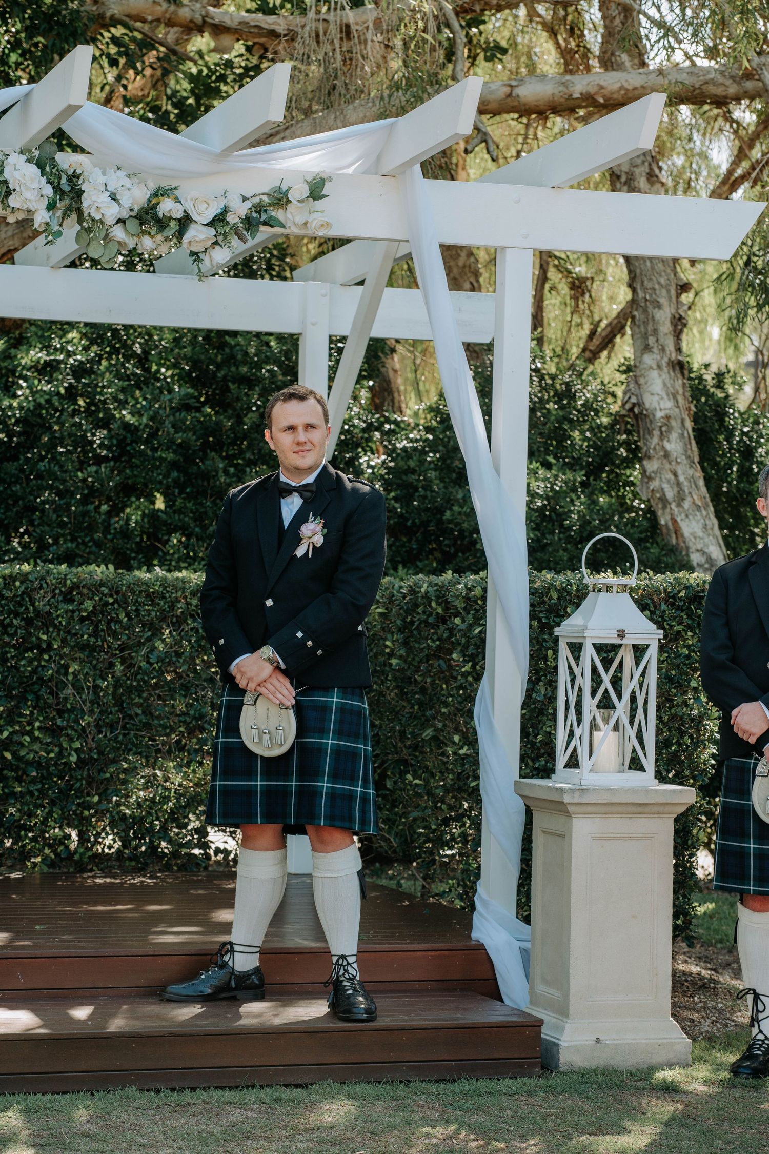 A person wearing a traditional Scottish kilt stands beneath a white pergola decorated with fabric draping in an outdoor setting.
