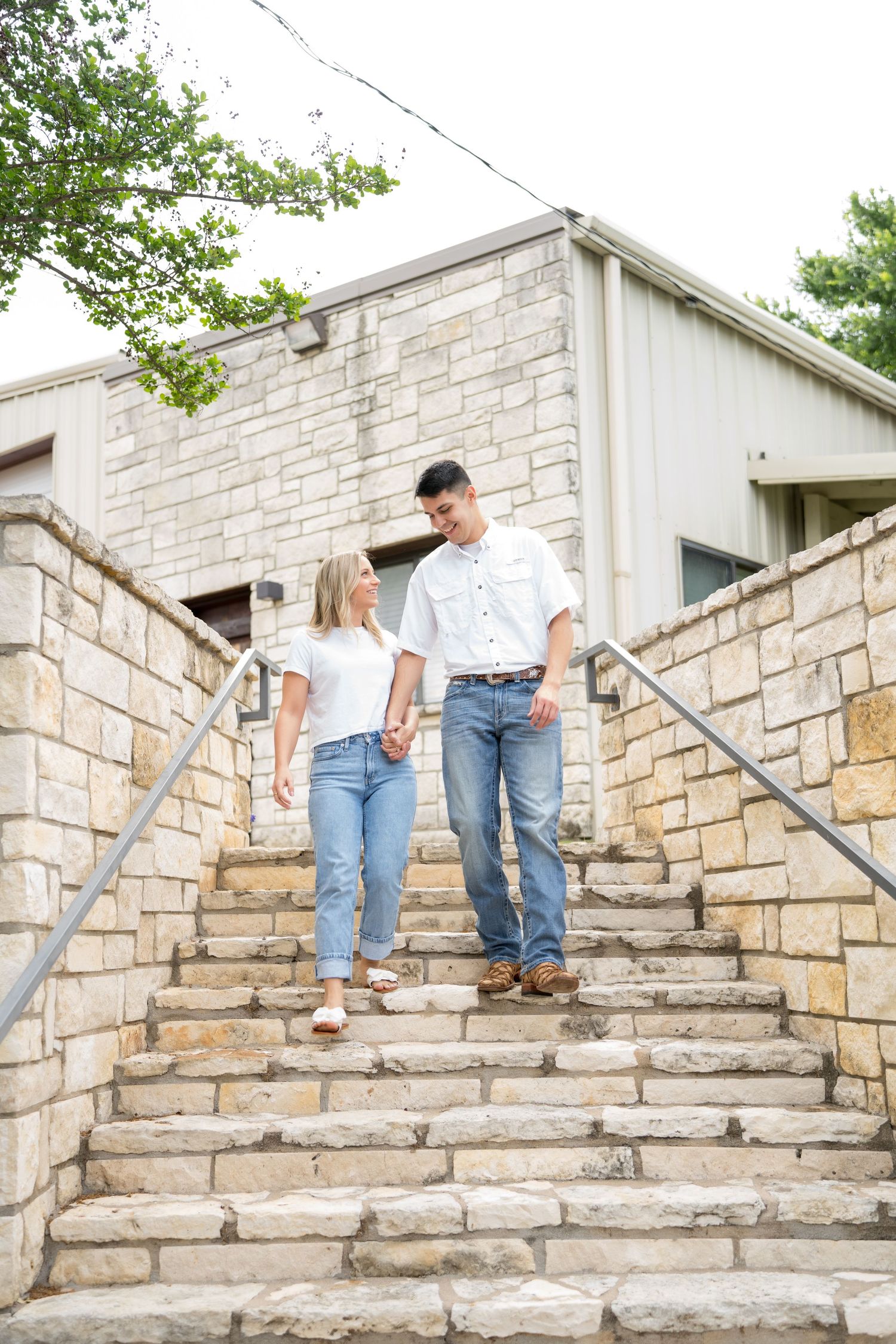 Couple holding hands walking down stone steps outside a rustic building, wearing matching white shirts and jeans.