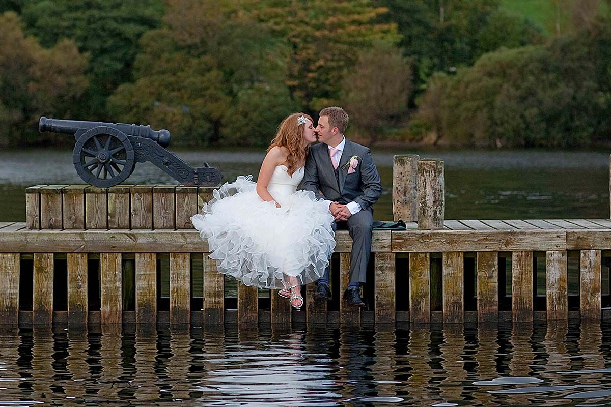 A romantic wedding couple sits on a wooden pier at low wood bay beside lake windermere, the lake district