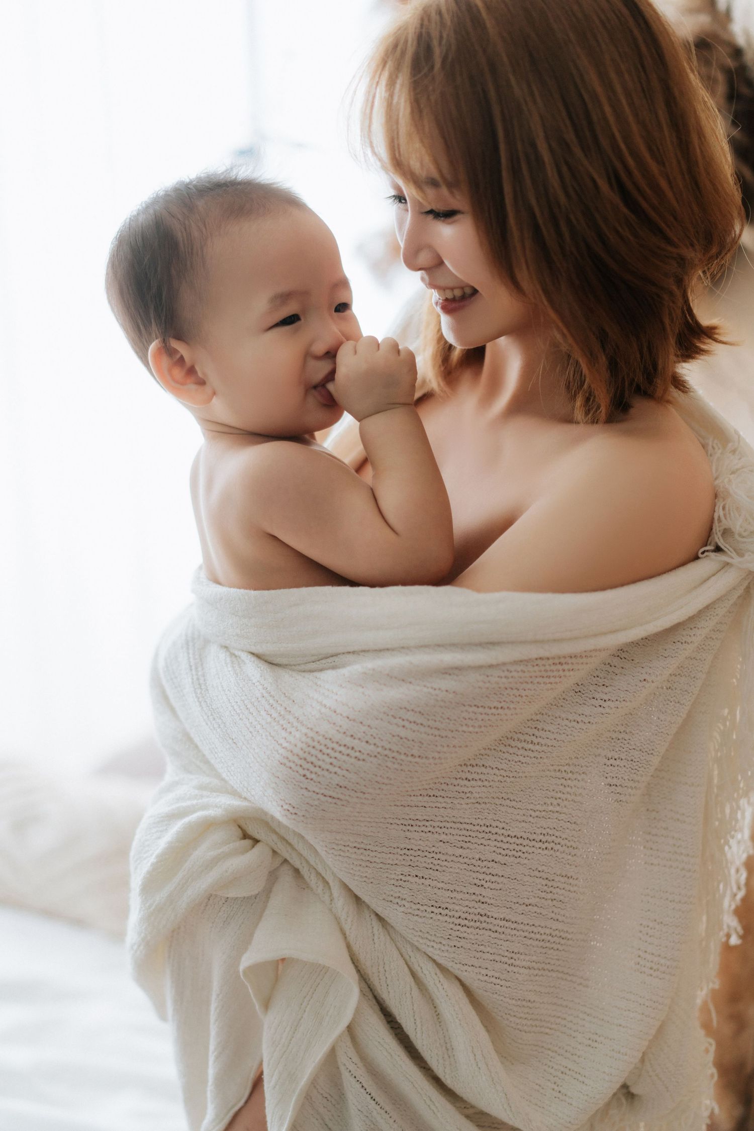 A tender moment between parent and baby wearing white, captured in soft natural light against a bright background.在柔和的自然光下，明亮的背景襯托著身穿白色衣服的父母