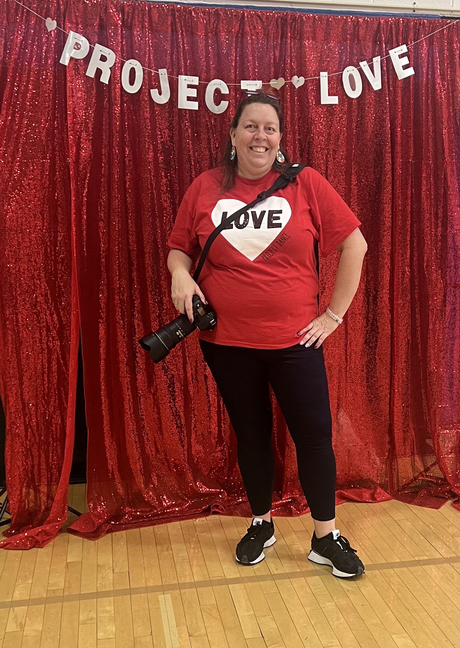 A photographer in a red LOVE t-shirt stands against a sparkly red backdrop with 'PROJECT LOVE' banner overhead.