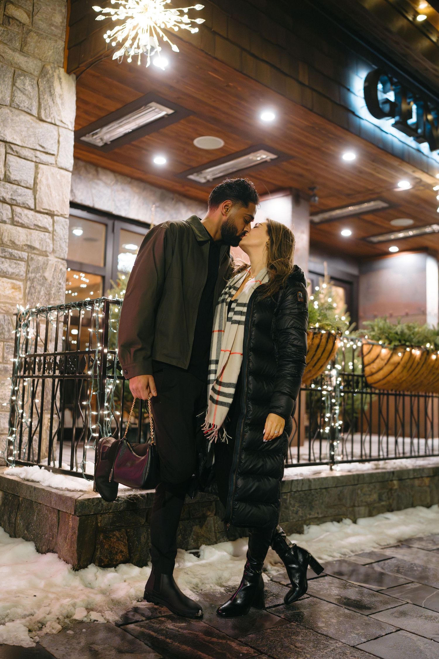 A couple shares an intimate moment outside a stone building with holiday lights and decorations in the evening.