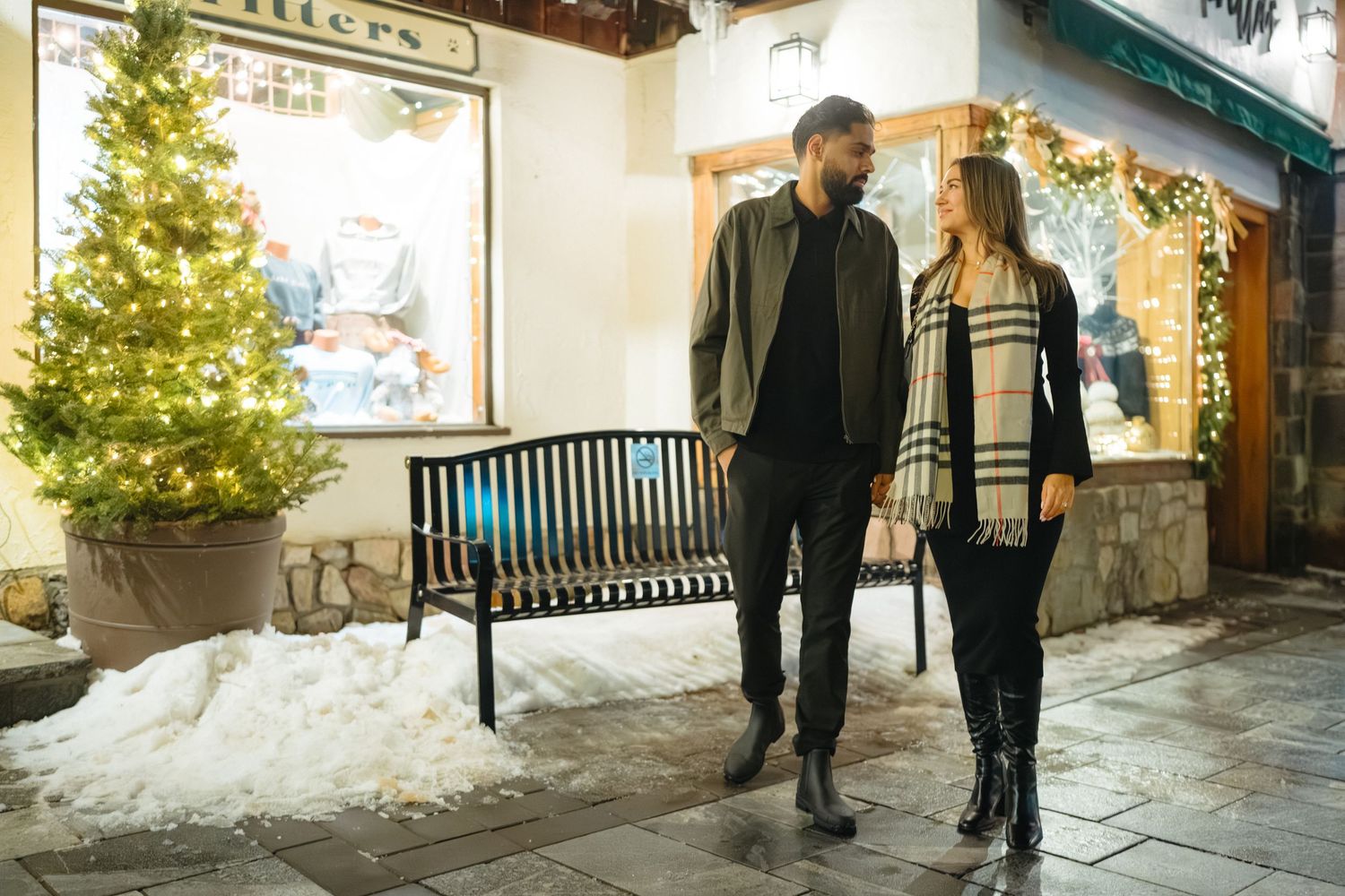 Couple strolling past decorated holiday storefront with lit Christmas tree and snow on ground.