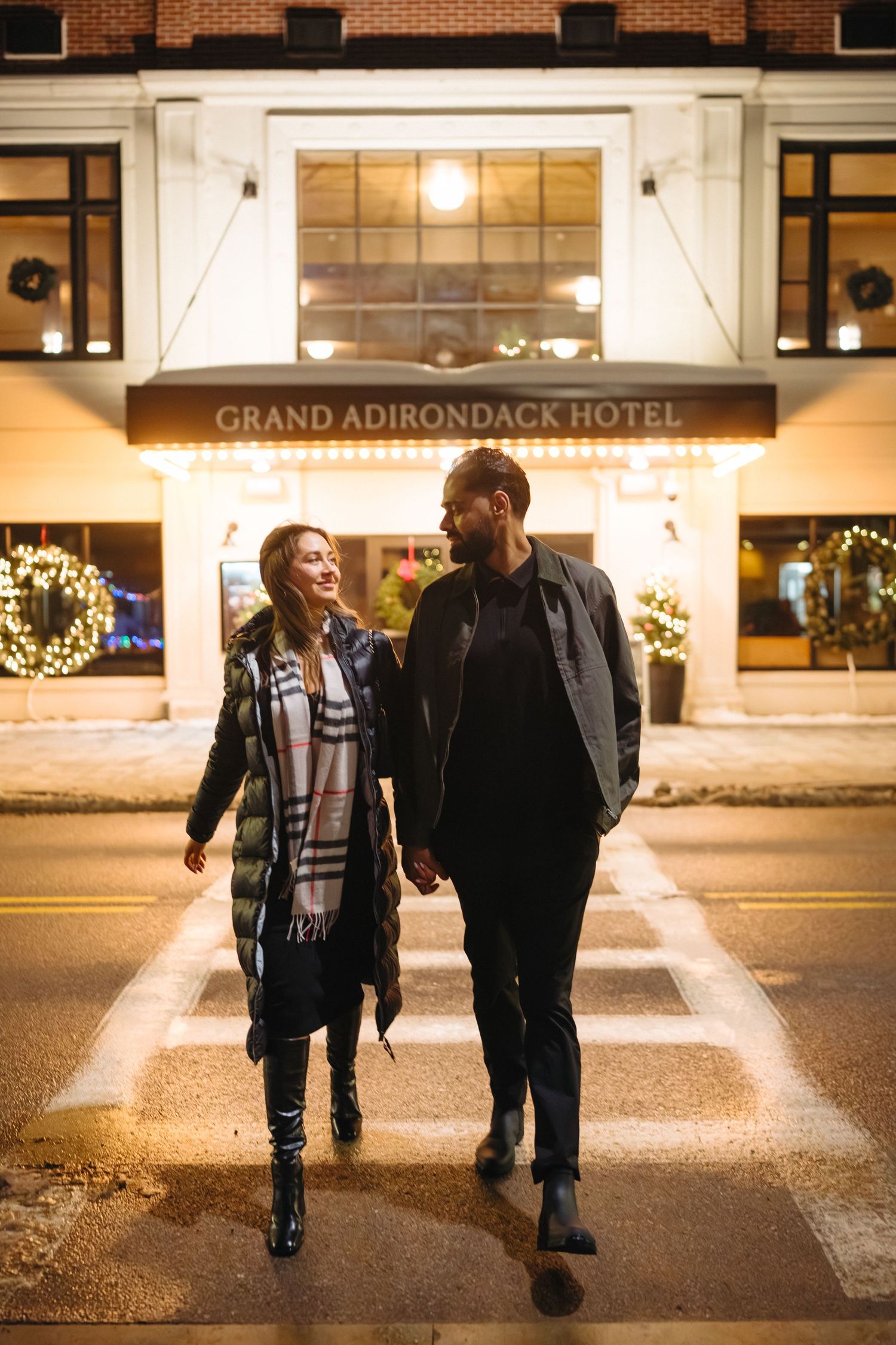 A couple walking together at night outside the Grand Adirondack Hotel with festive holiday lights in the background.