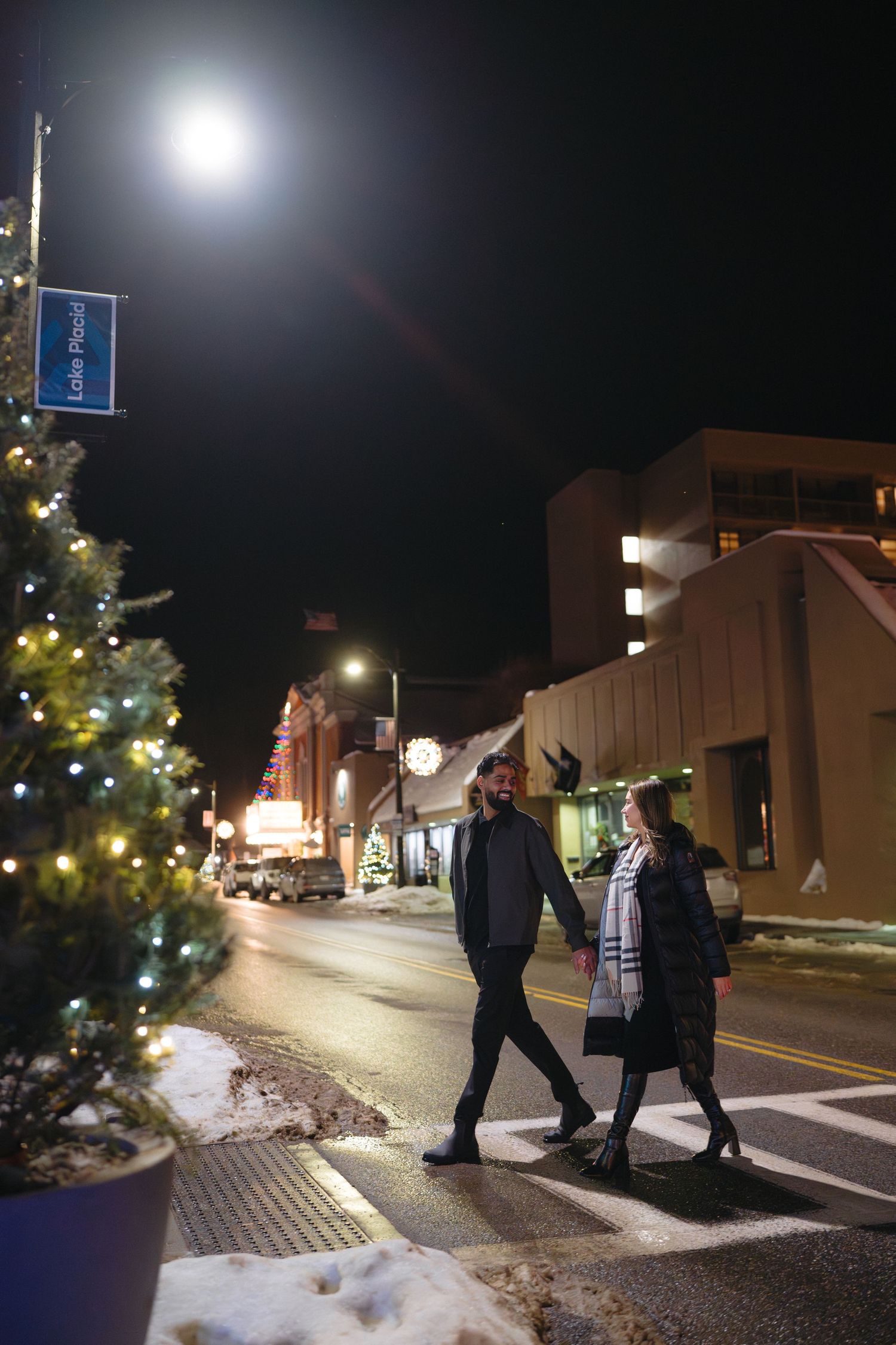 Couple walking together at night on a snowy small town street with lit Christmas trees.