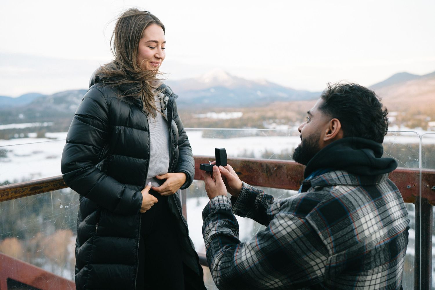 A couple shares tender moments on a balcony overlooking a scenic mountain landscape during winter.