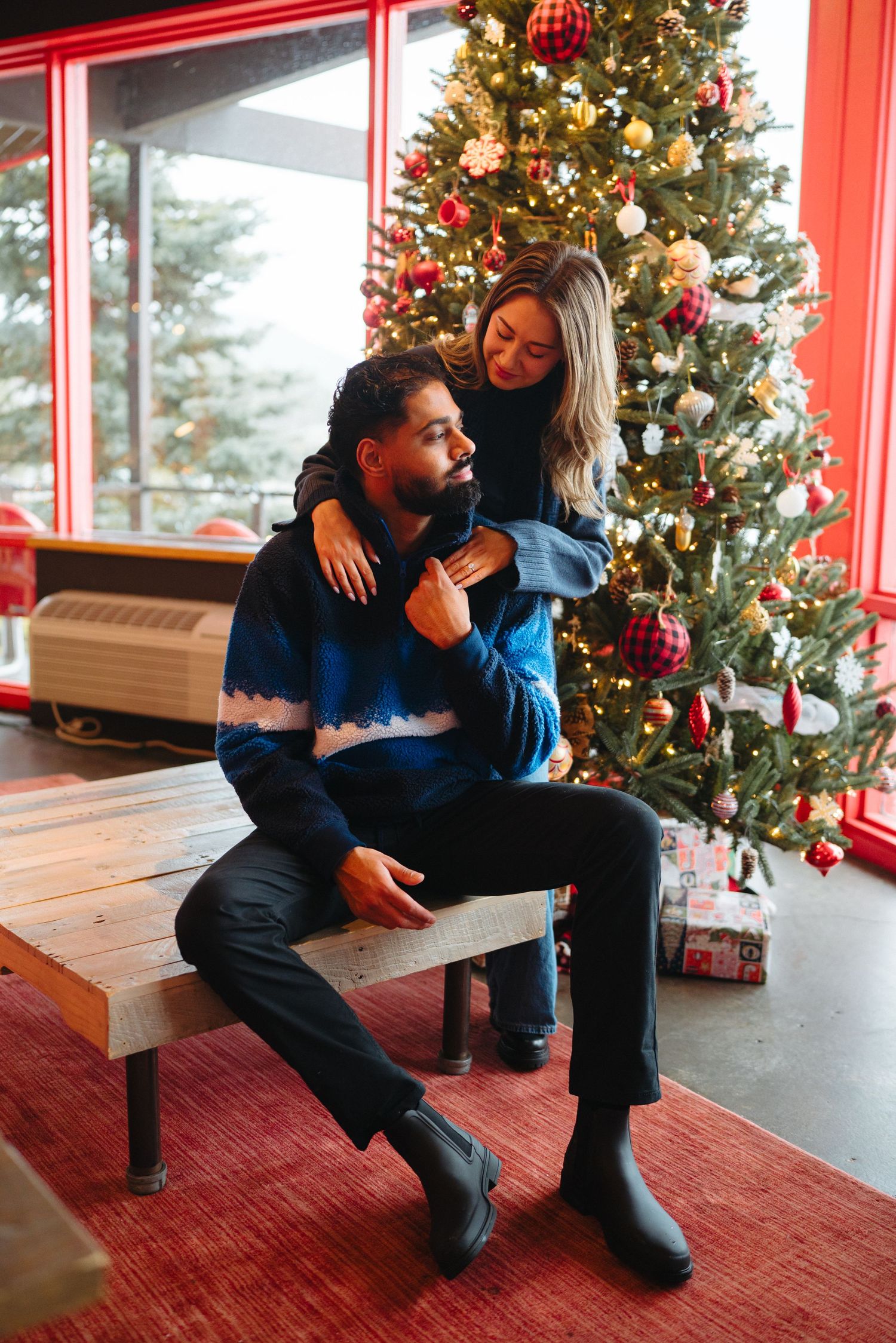 A couple shares a tender moment by a decorated Christmas tree with wrapped presents underneath.