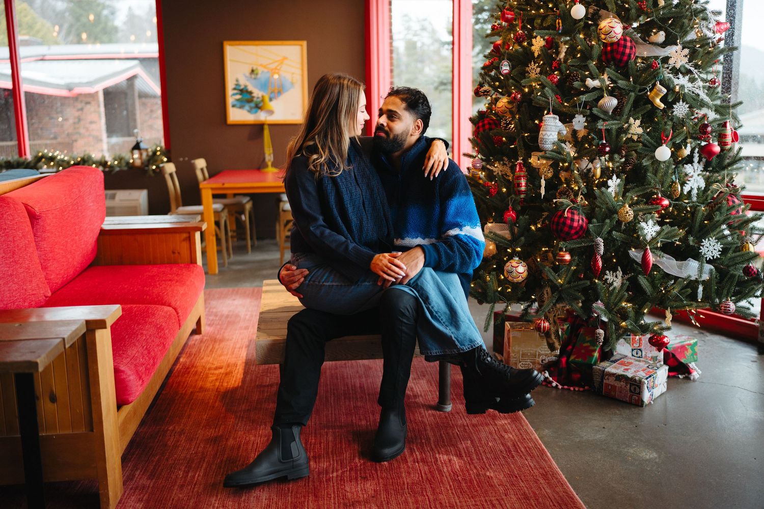 A couple sits together on a red couch near a decorated Christmas tree in a cozy living room setting.