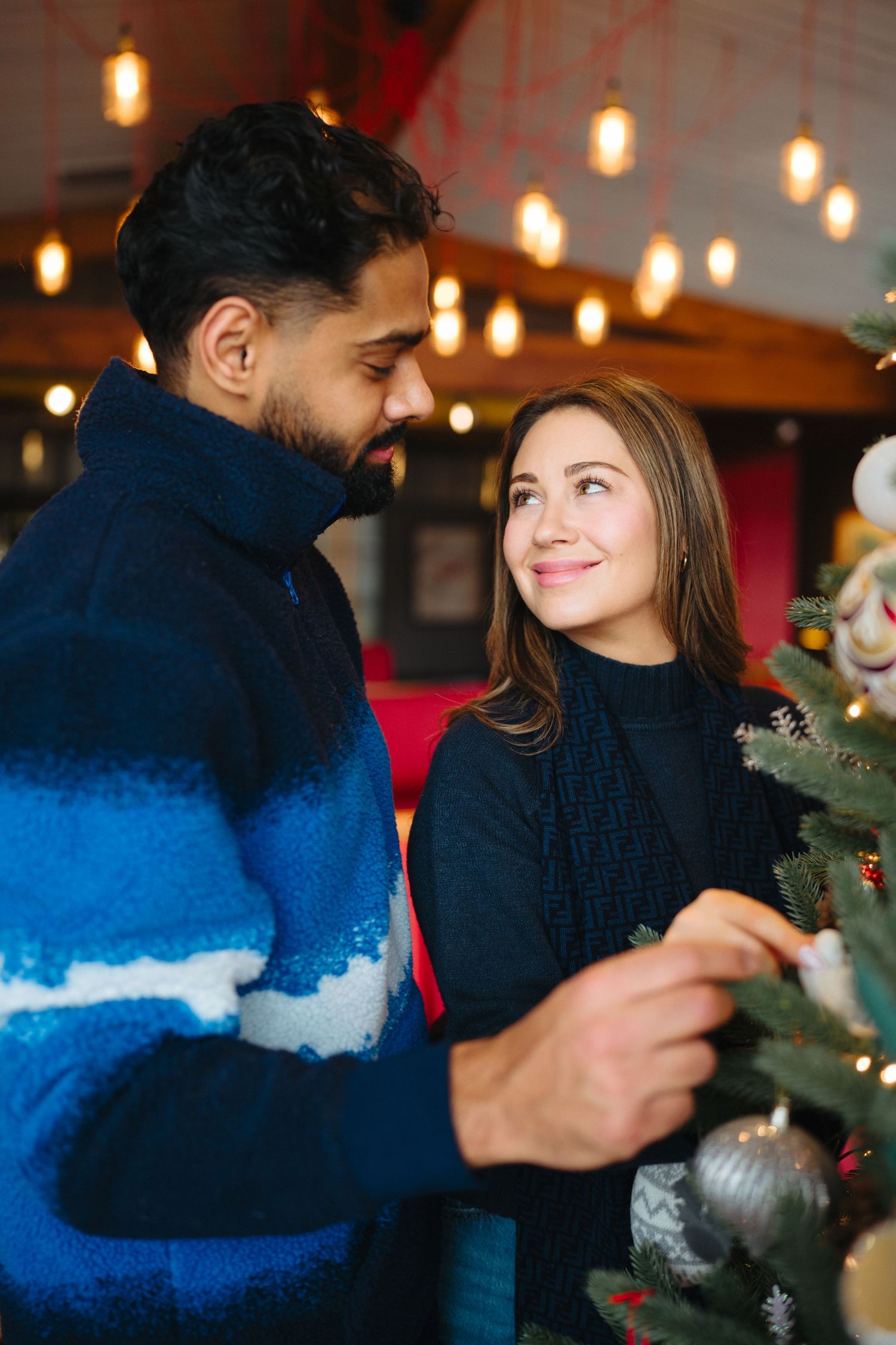 A couple decorates a Christmas tree together in a warmly lit room with hanging pendant lights.