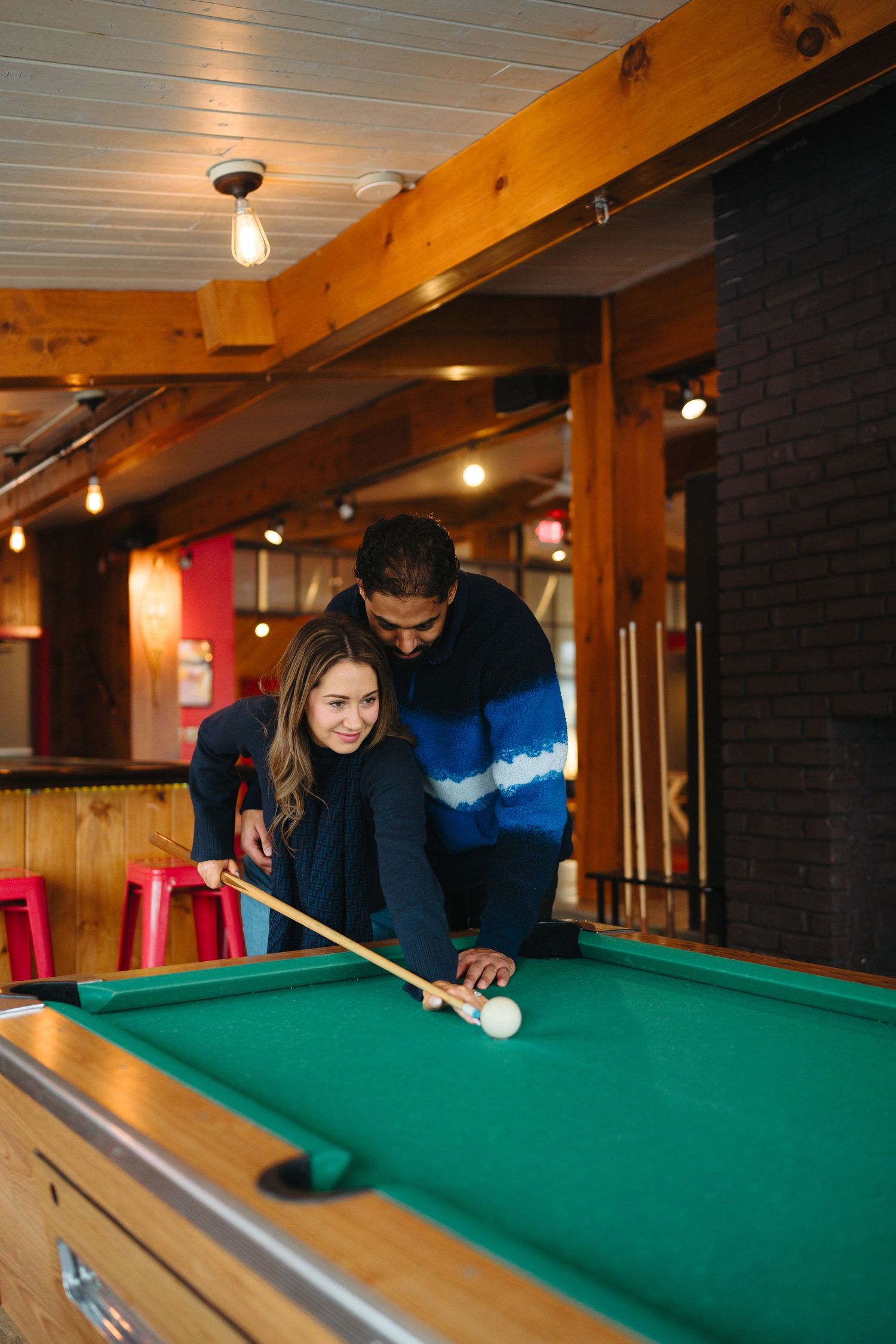A couple playing pool together in a rustic room with wooden beams and ambient lighting.