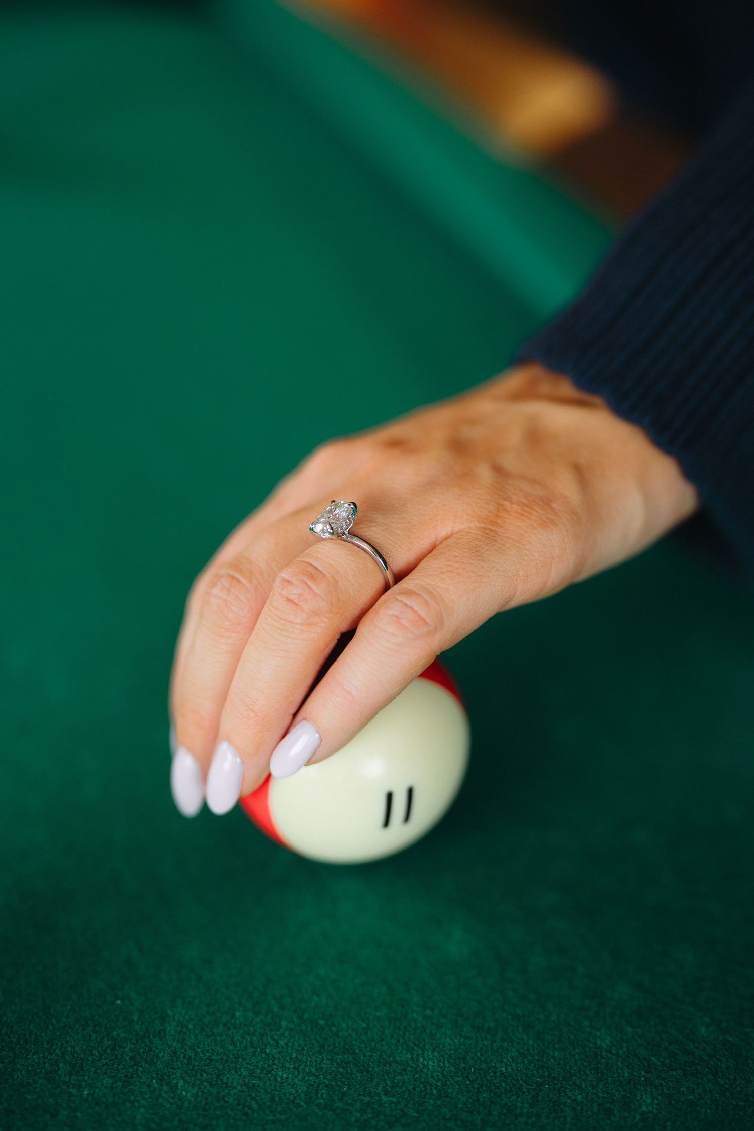 A hand wearing a diamond engagement ring rests on a white billiard ball on a green felt pool table.