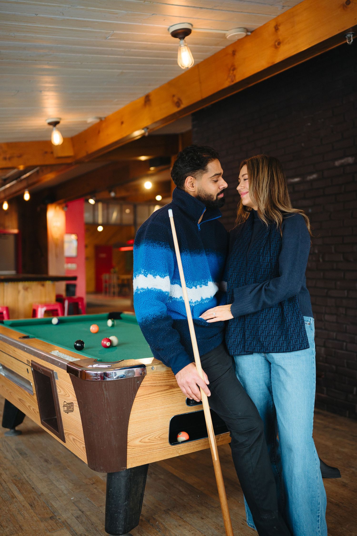 Couple sharing a tender moment in a rustic pool hall with wooden beams and billiard tables.