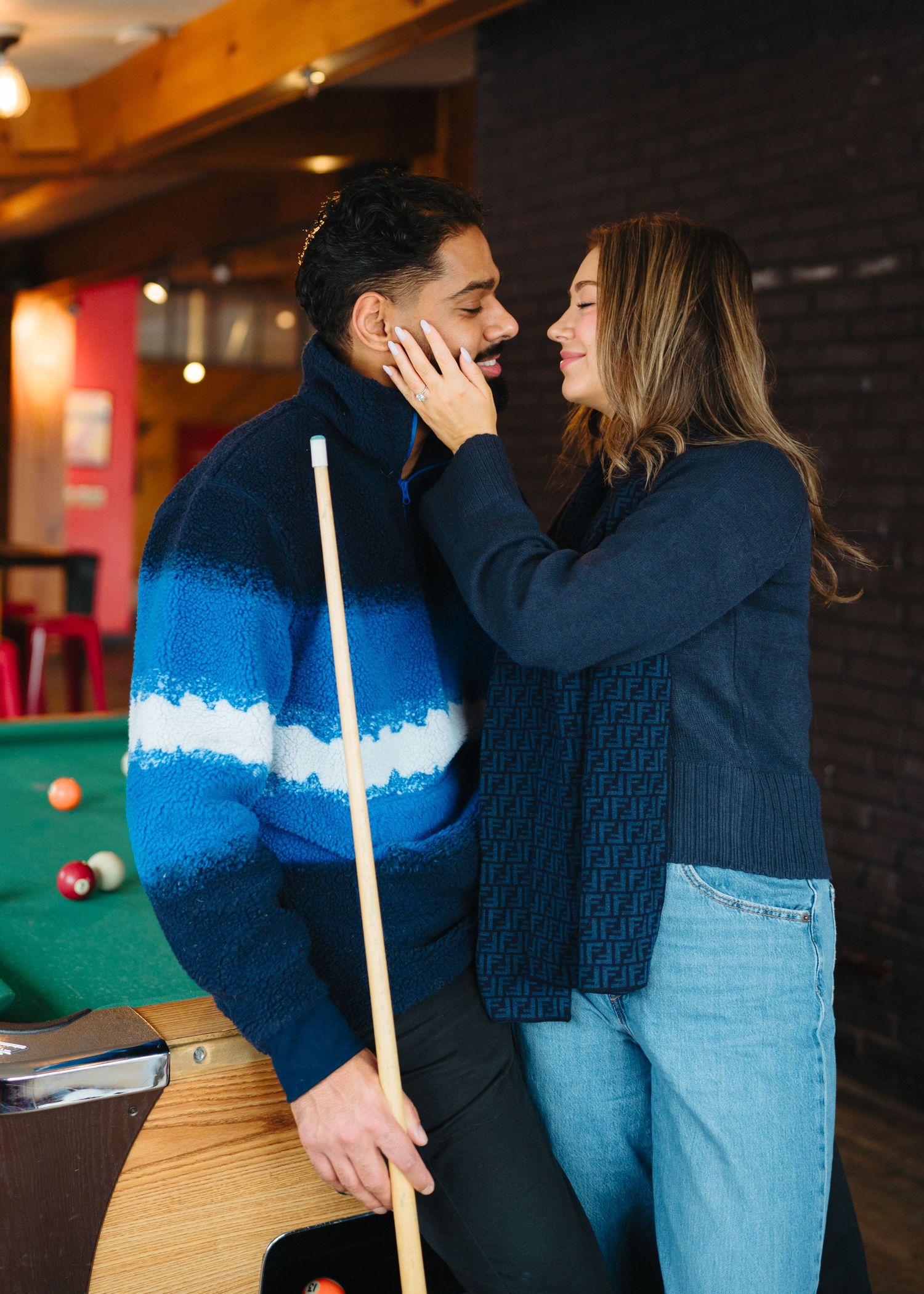 A couple shares an intimate moment while playing pool, with one holding a cue stick and wearing a blue sweater.