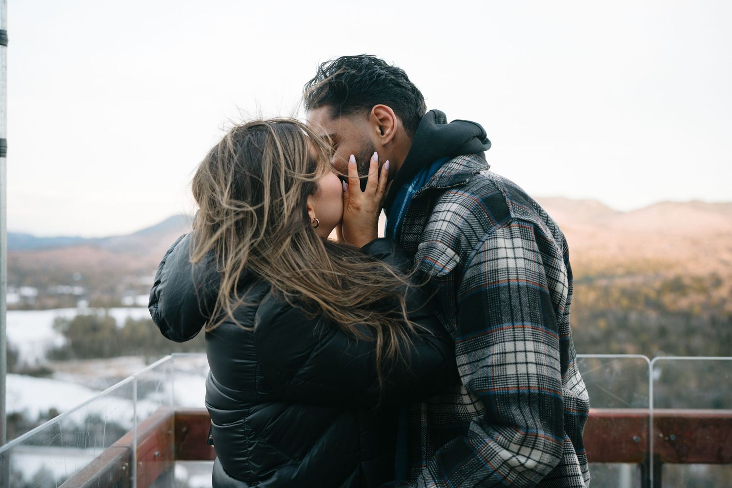 Two people embrace on a wooden deck with scenic mountain views in the background.