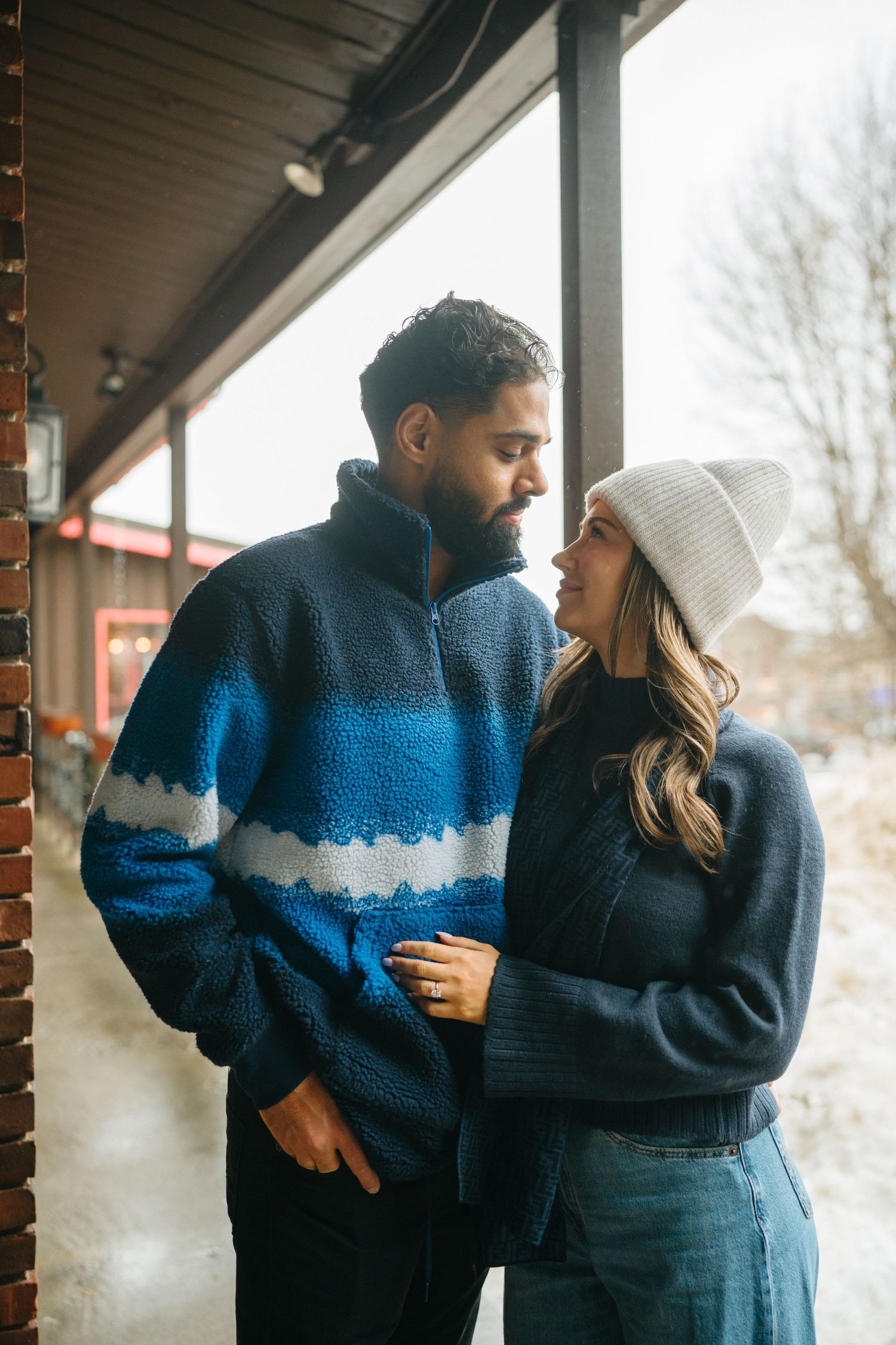 A couple shares an intimate moment on a porch wearing cozy winter sweaters and a white beanie.