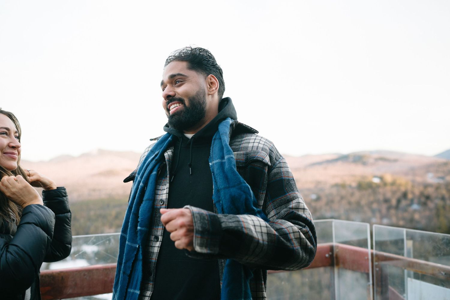 Someone laughs while wearing a blue denim jacket and plaid coat on an outdoor deck with mountains in the background.