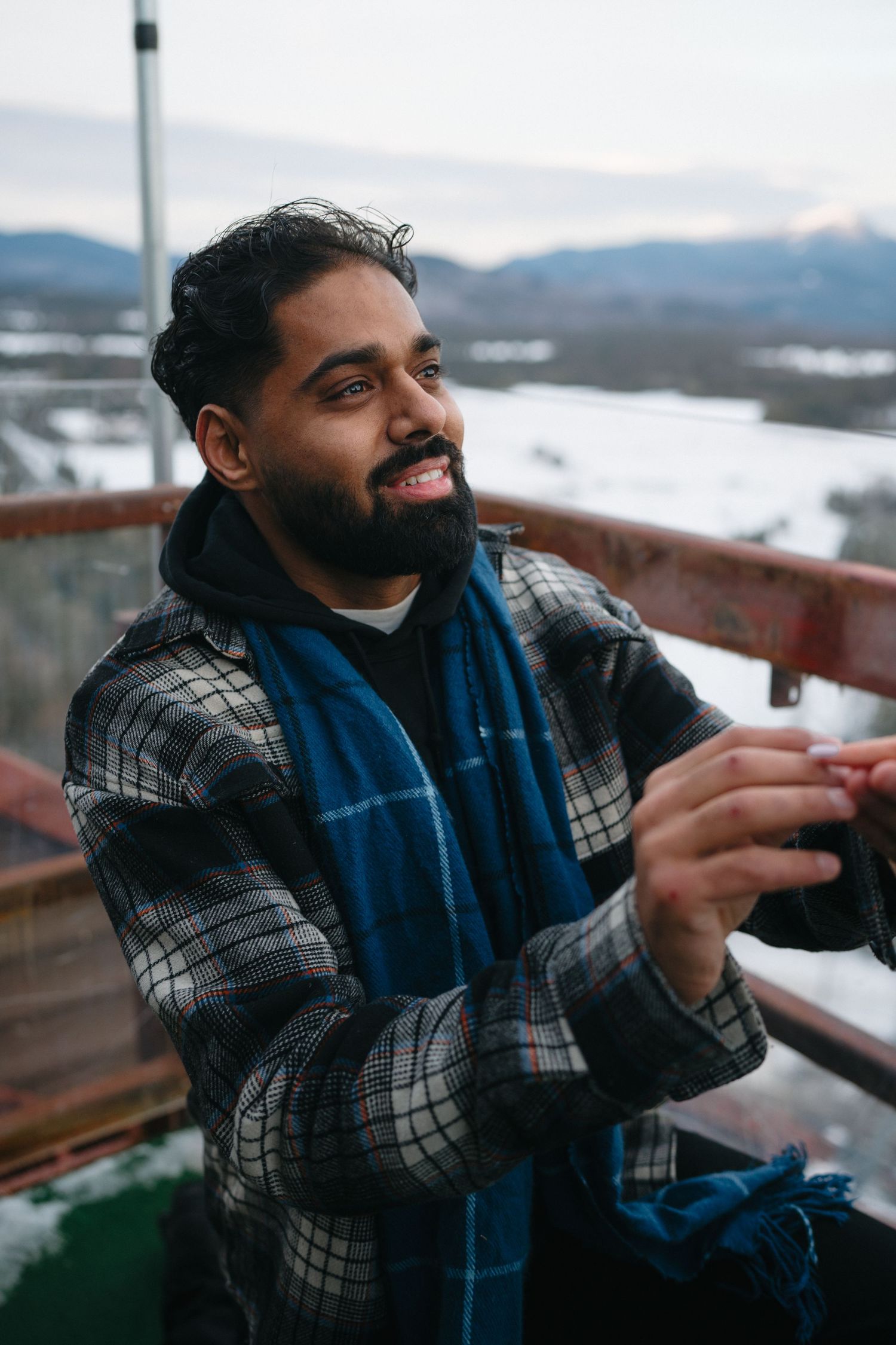 Person in plaid jacket and blue scarf gesturing while enjoying mountain views from observation deck.