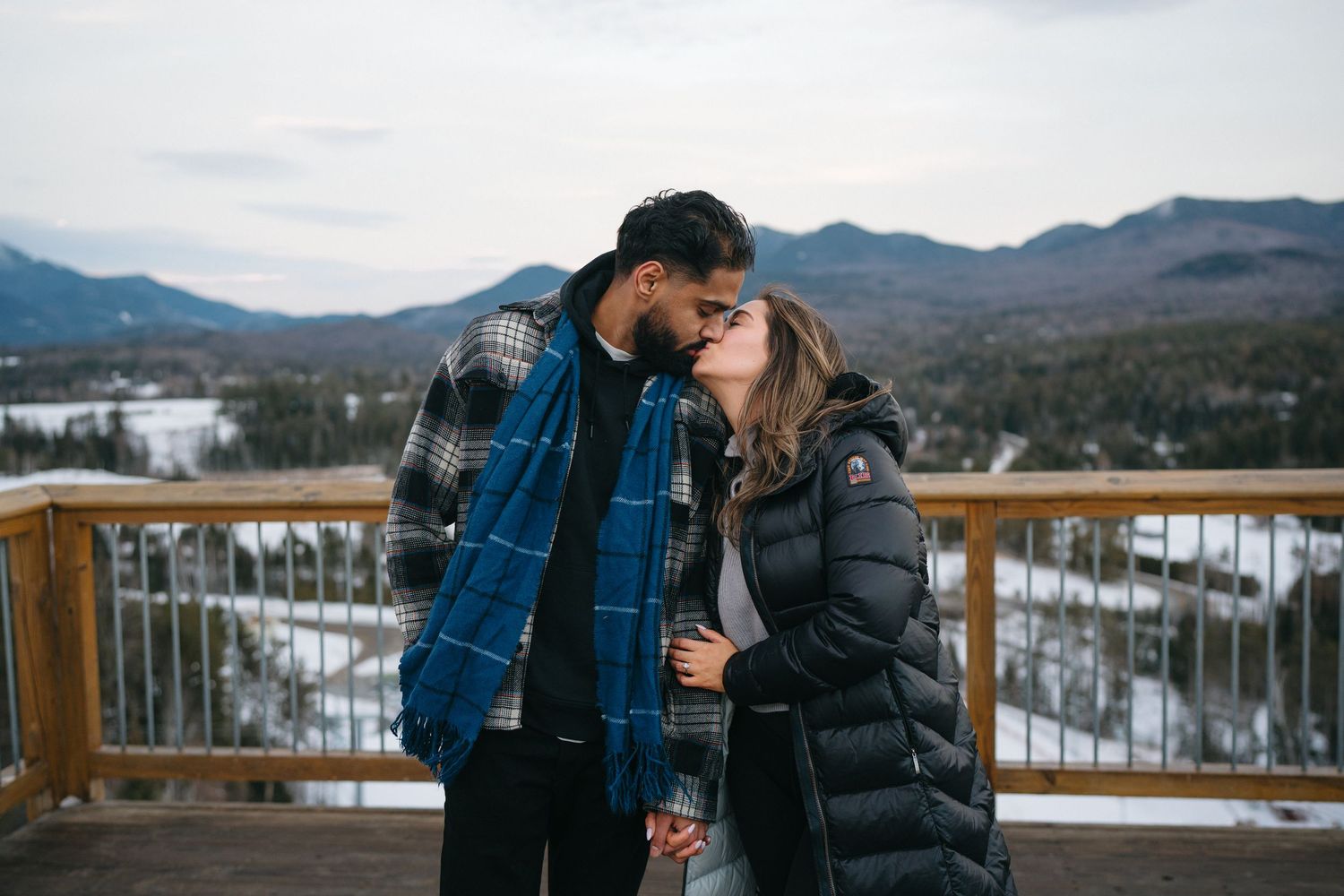 Couple embracing on a snowy mountain deck with scenic winter landscape views in the background.