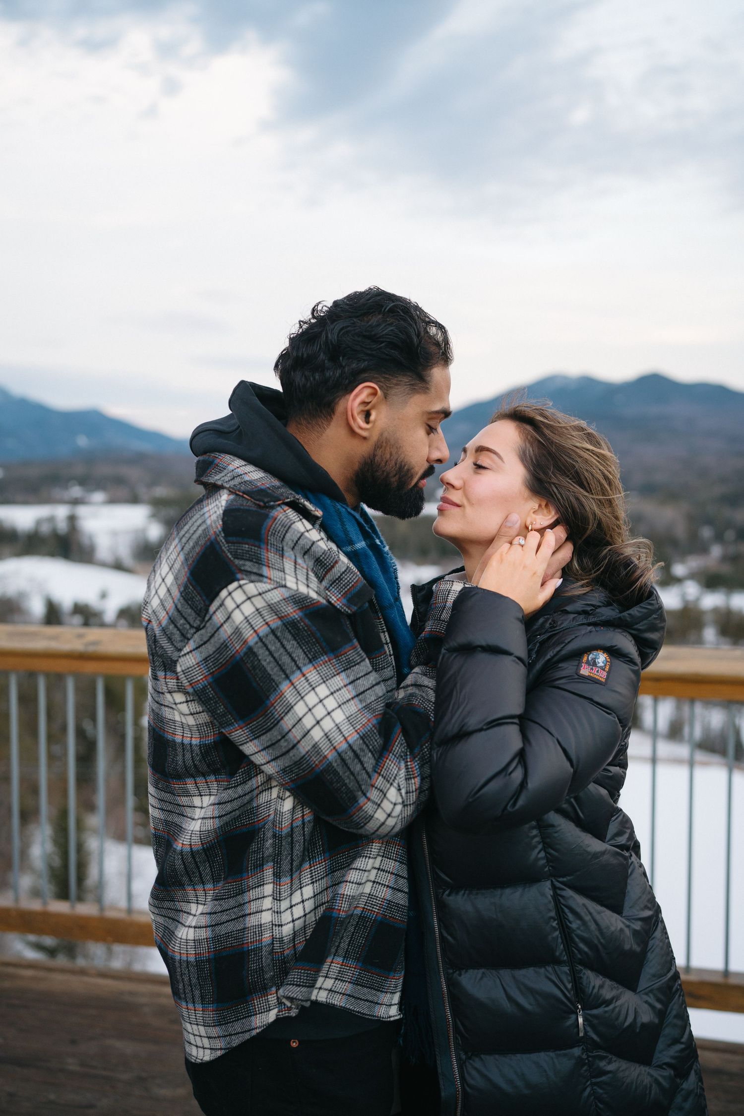 A romantic couple embraces on a deck overlooking snowy mountains, with one wearing a plaid flannel and the other in a puffy coat.