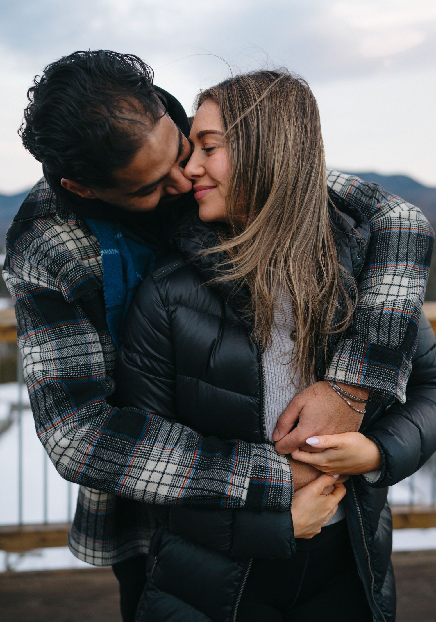 A romantic couple sharing intimate kisses outdoors in winter, wearing cozy plaid and black jackets against a mountain backdrop.