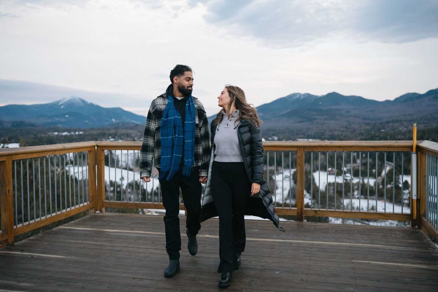Couple walking together on observation deck with snow-covered mountains in background.