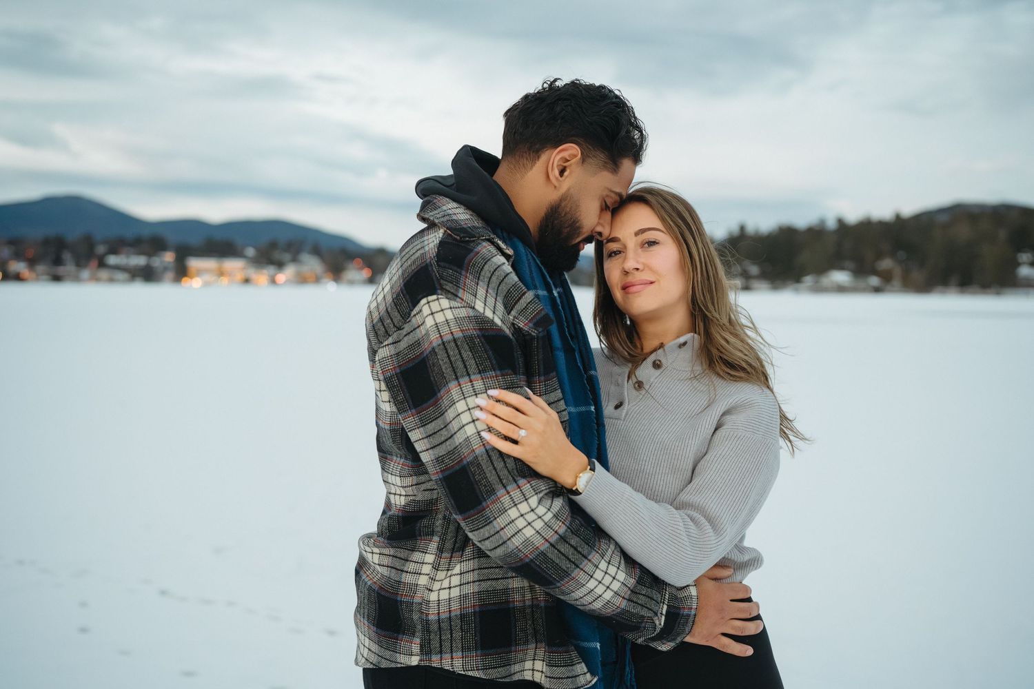 Couple sharing an intimate moment outdoors in winter with mountains and cloudy sky in background.