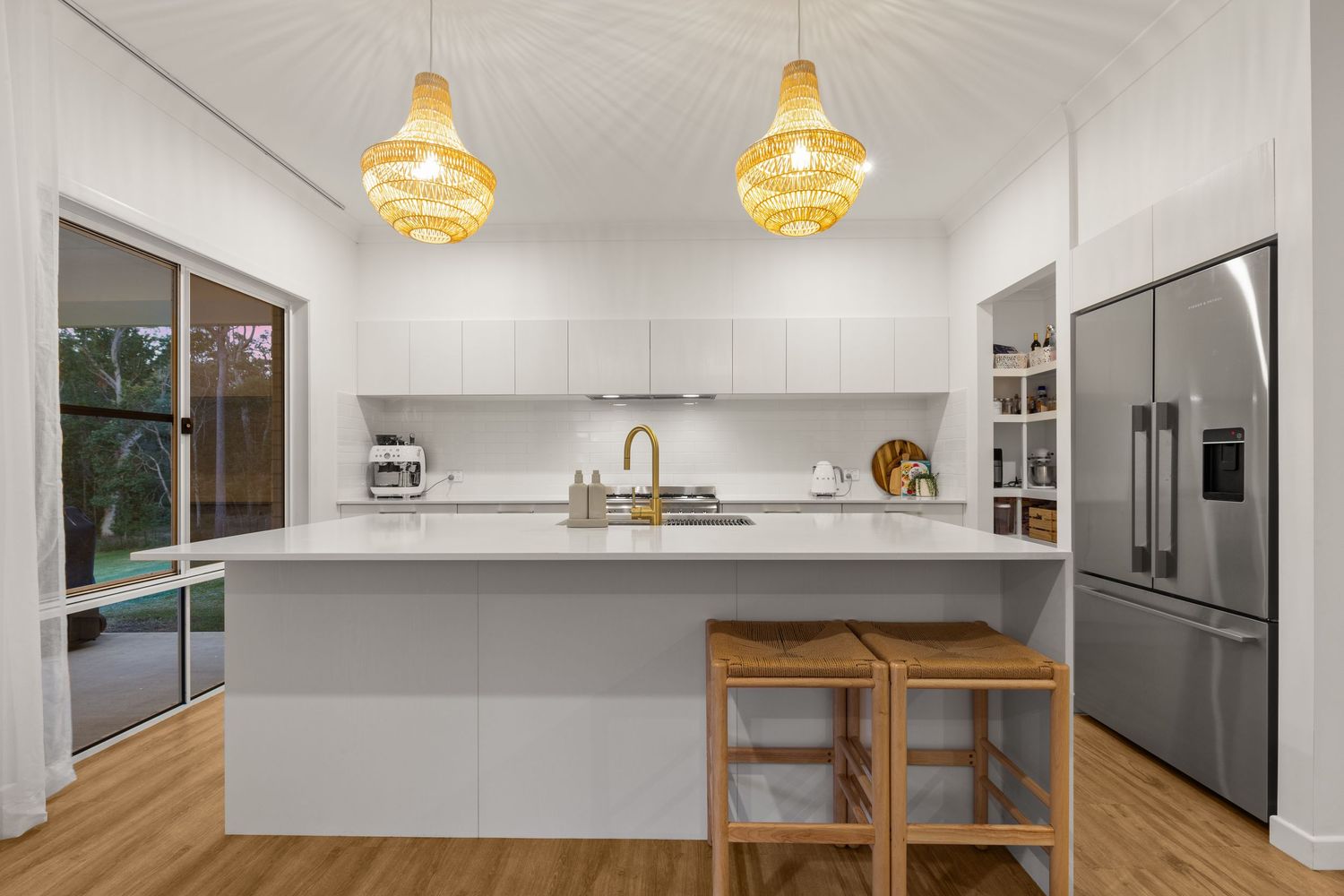 Modern white kitchen with island bench, timber bar stools, rattan pendant lights, and stainless fridge.