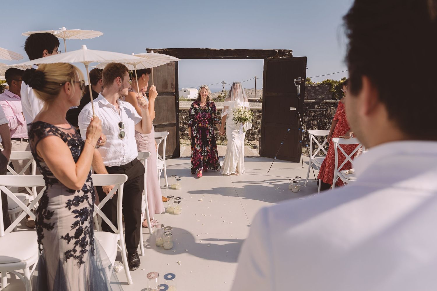 Outdoor destination wedding ceremony with guests standing as the bride enters El Viento in Santorini.