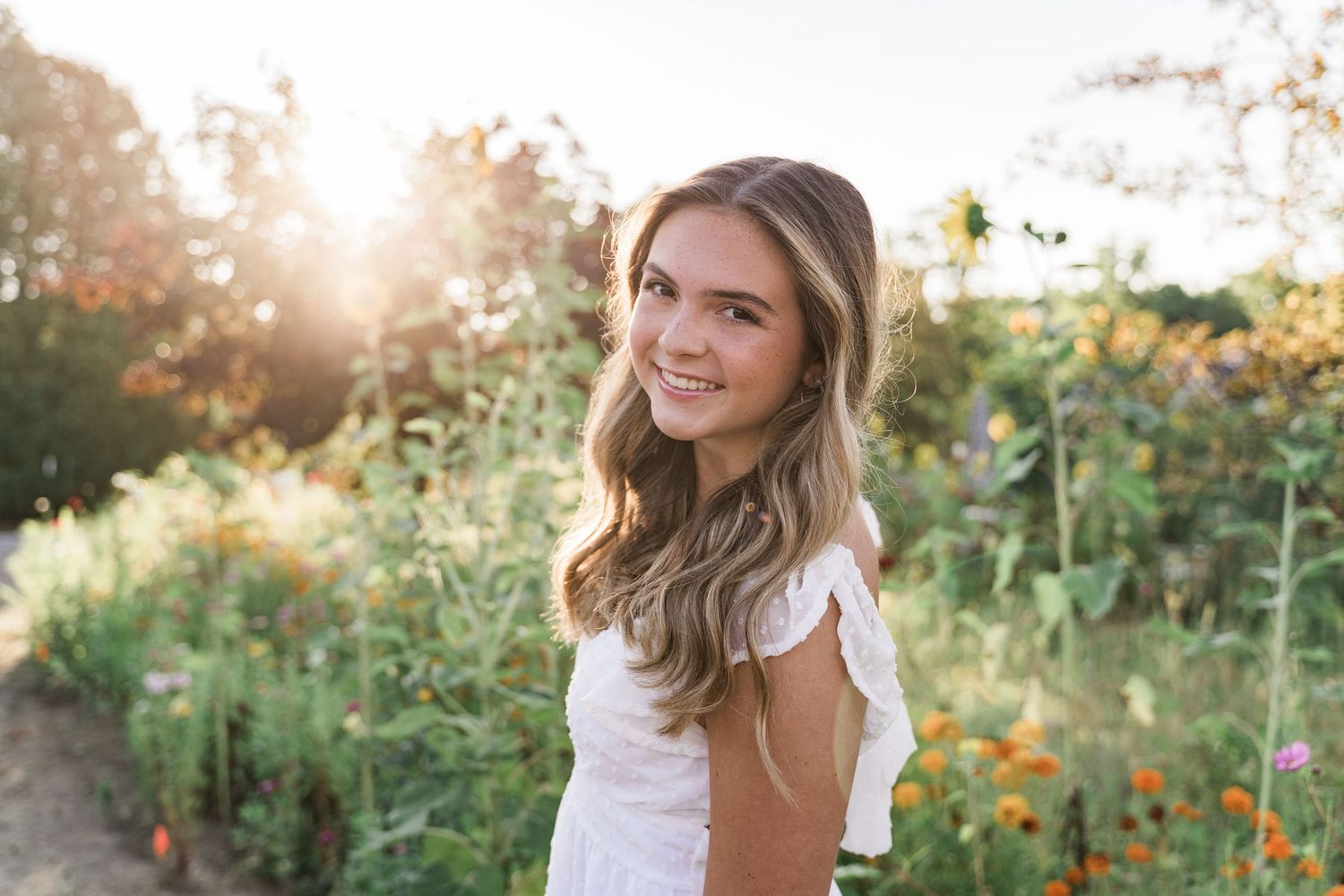 Senior in white dress poses in a sunlit garden with blooming flowers during golden hour sunrise at GardenView Flowers in Grand Rapids, Ohio.