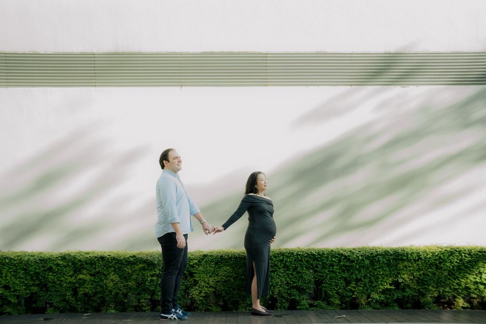 A couple holds hands while standing against a white wall with green hedges, creating a minimalist outdoor scene.