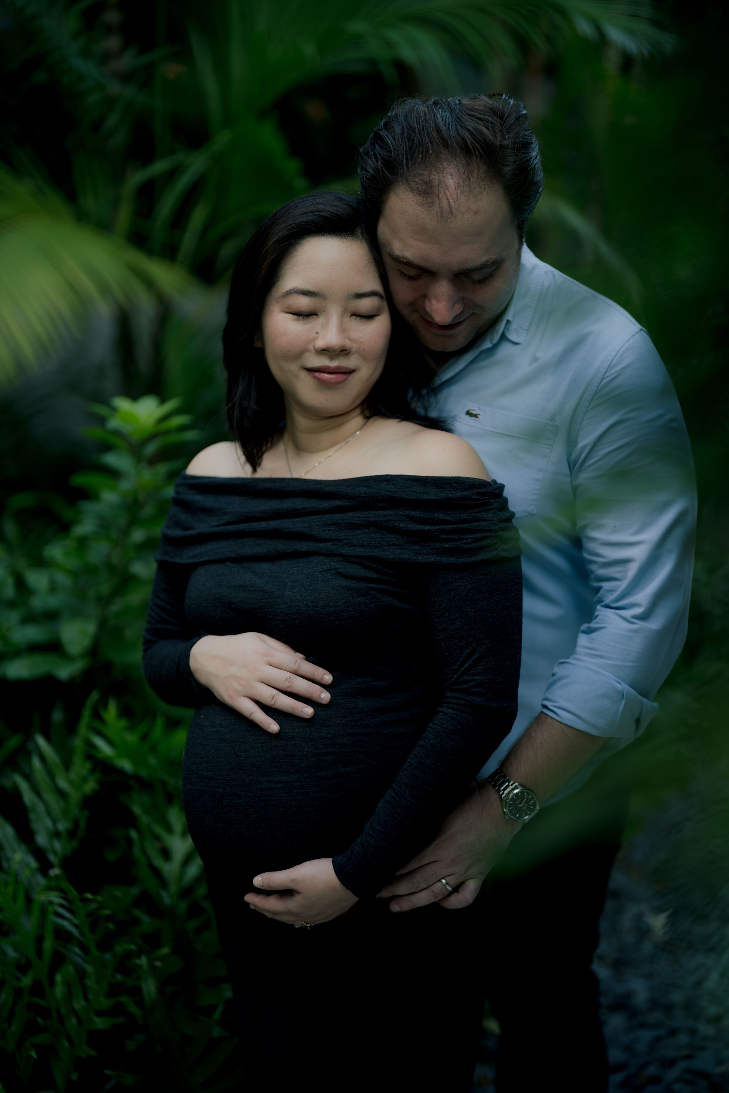 Intimate maternity photo series showing couple embracing in lush garden setting with black dress and natural lighting.