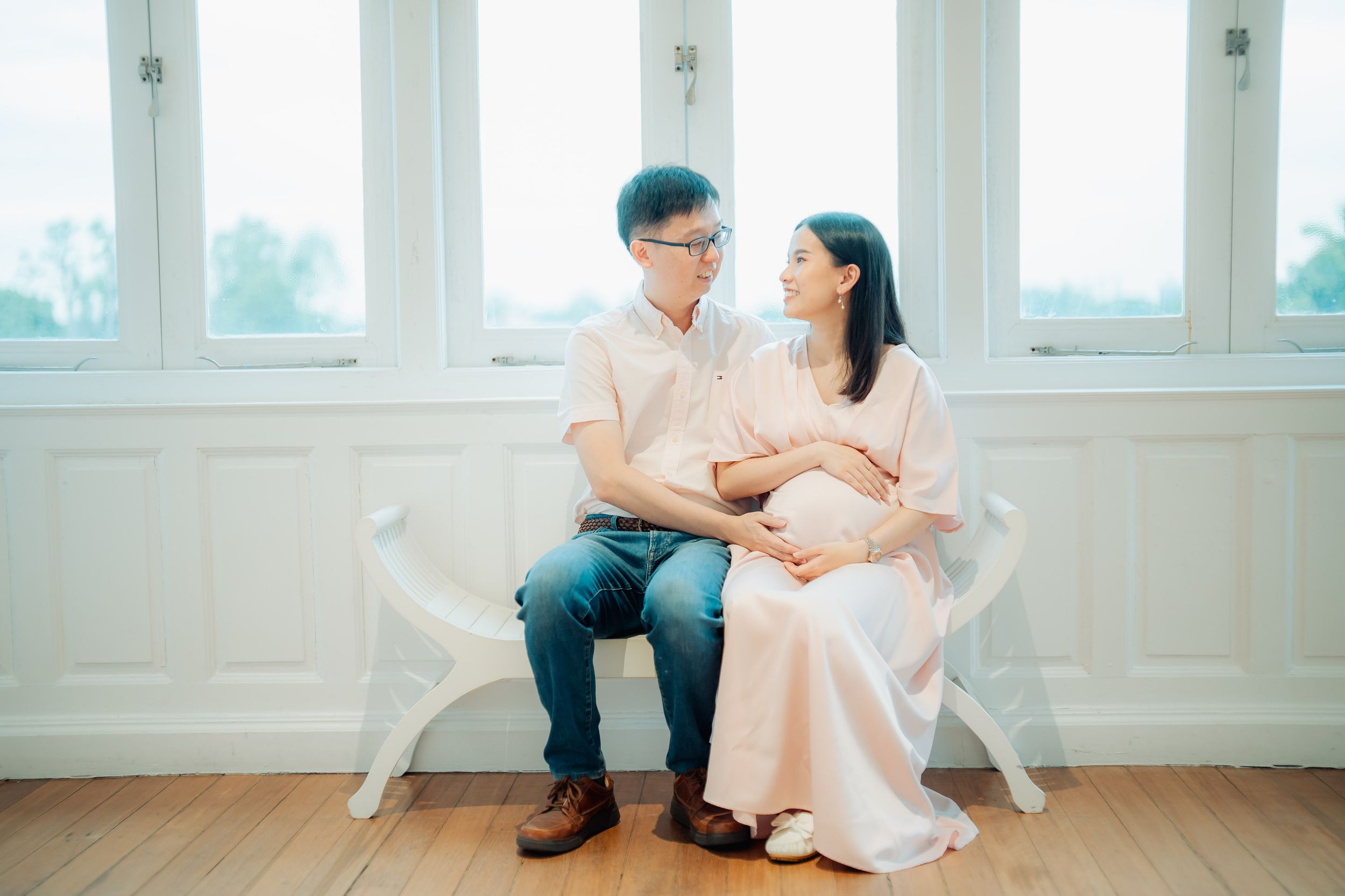 Couple sitting in white chairs by large windows in bright minimal interior setting.