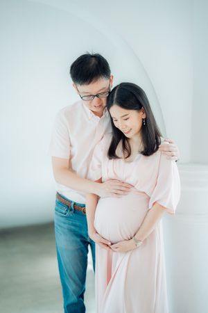 An expecting couple in matching pink outfits sharing a tender moment against a white backdrop.
