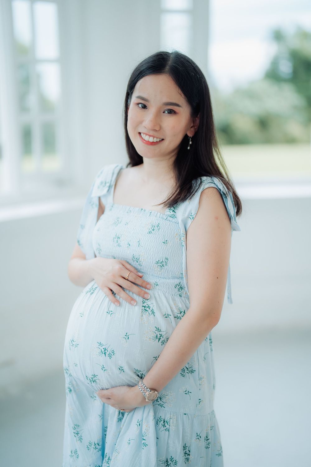 An expectant mother in a light blue floral dress poses by a bright window.