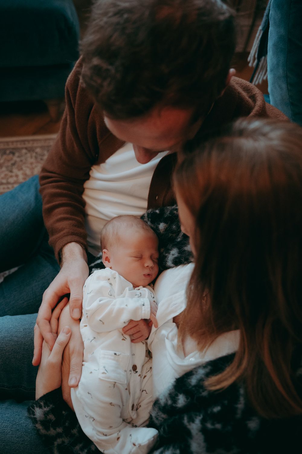 A sleeping newborn baby in white clothing rests peacefully between two people with a cozy home setting in the background.