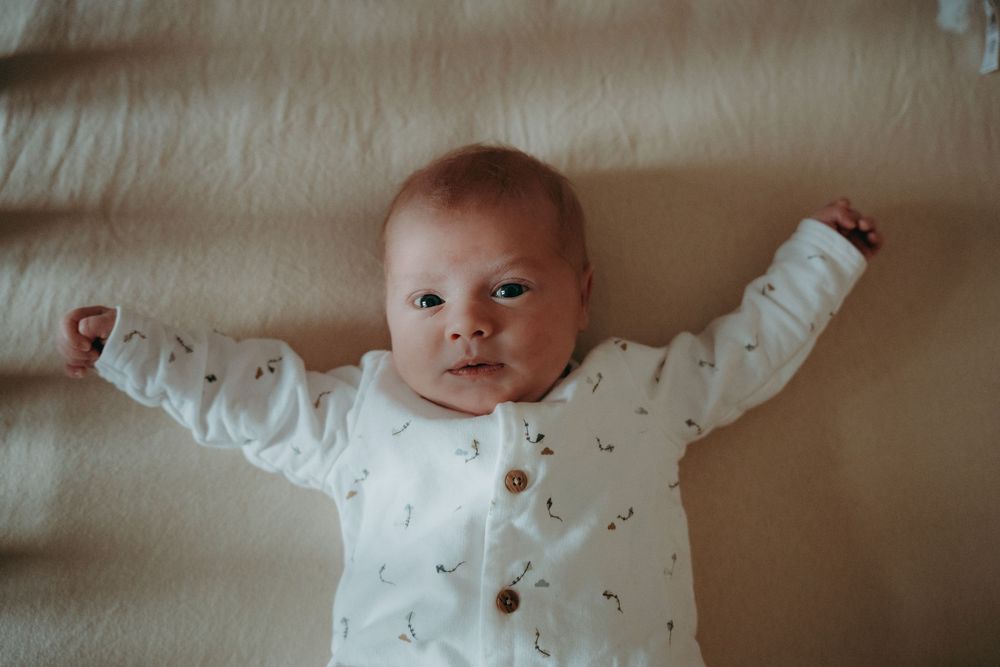 A baby wearing a white button-up outfit lies on beige bedding with arms stretched outward.