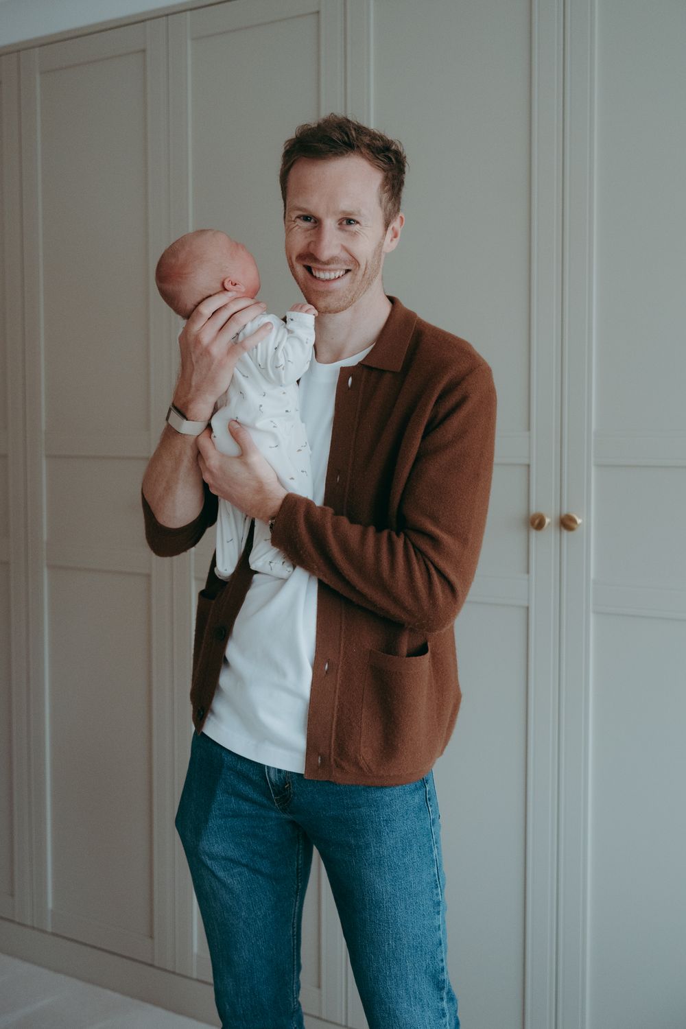 Person wearing brown cardigan and jeans holds and plays with small baby in white outfit against white door.