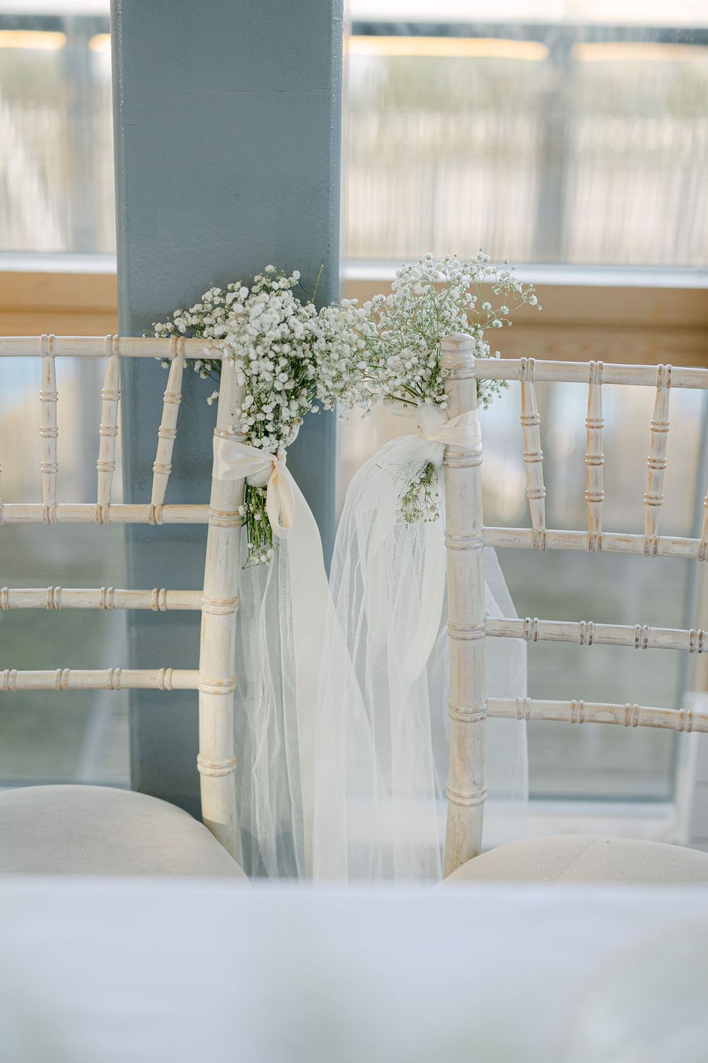 White chiavari chairs adorned with delicate baby's breath flowers and tulle decorations at a wedding ceremony.