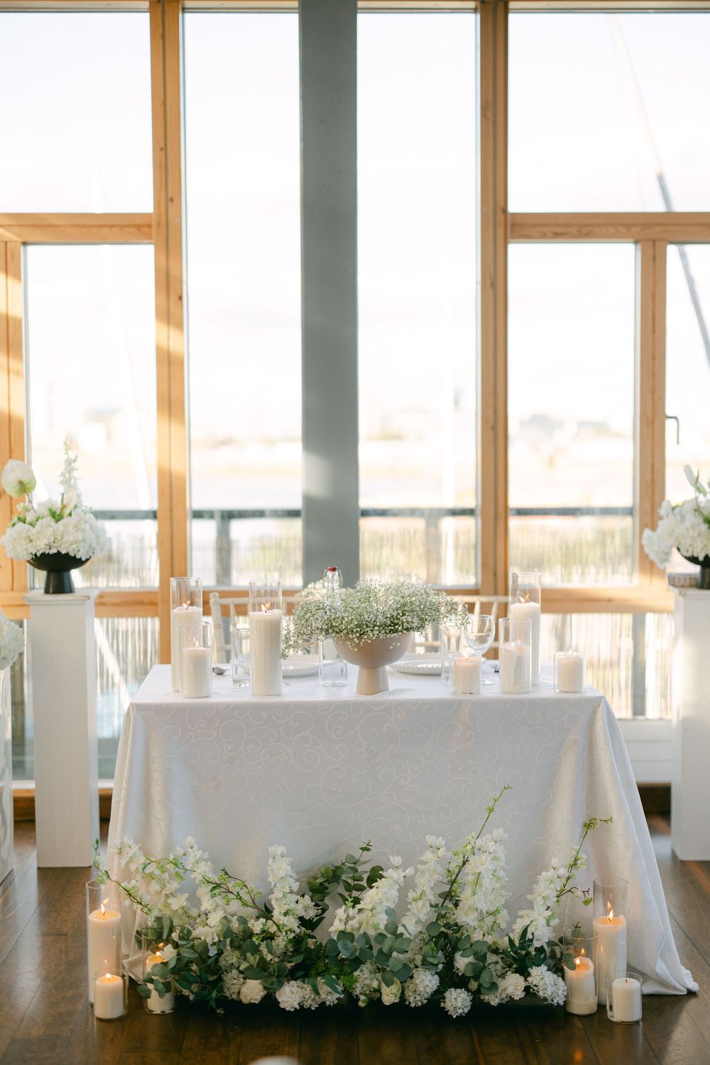 Bright sunlit wedding reception venue with white-clothed tables and floral arrangements by large windows.