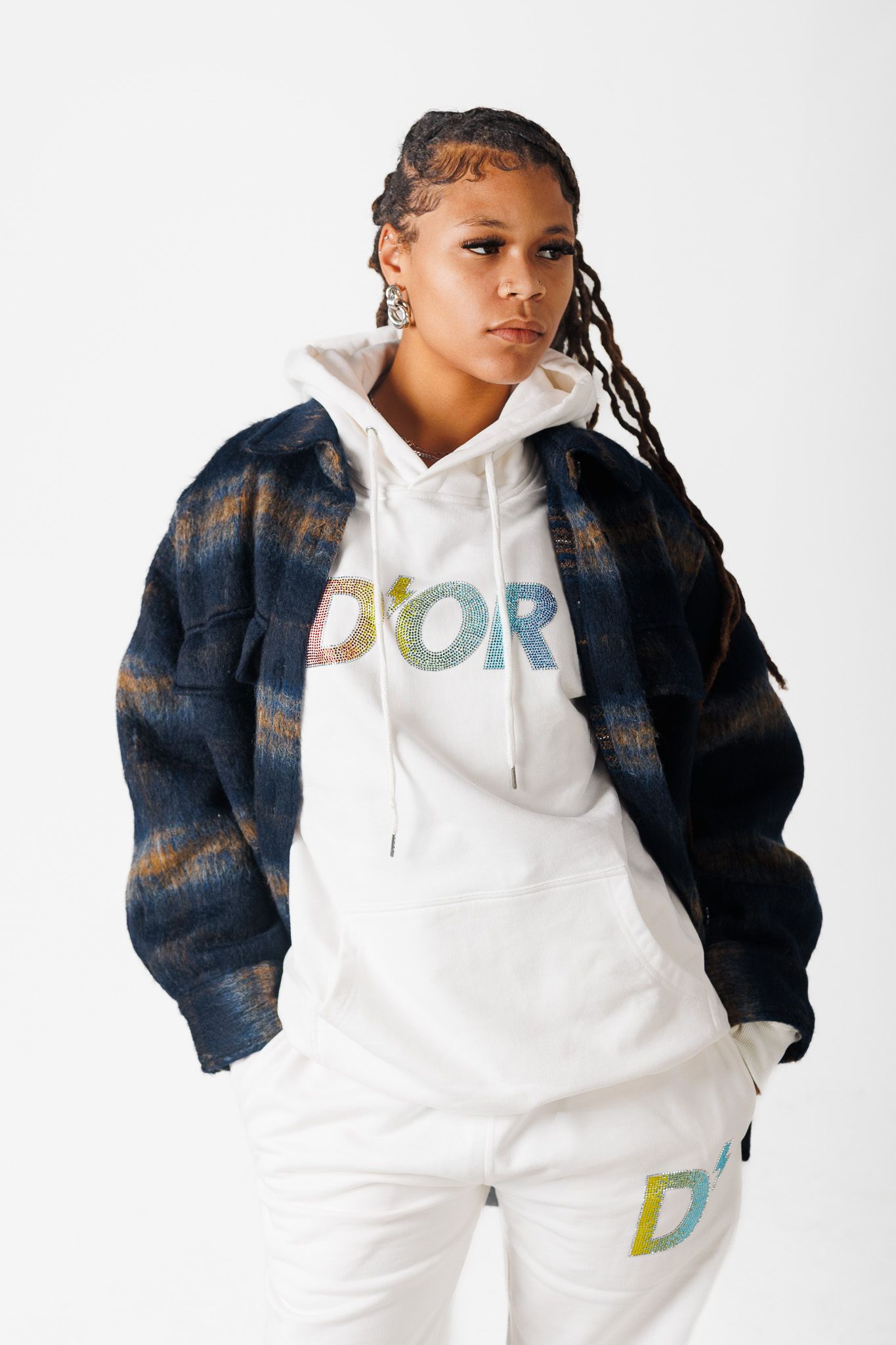 Person wearing white hoodie and dark patterned jacket poses on white background showing casual streetwear style.