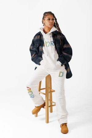 Model wearing white overalls and denim jacket poses on wooden stool against white background in fashion photoshoot.
